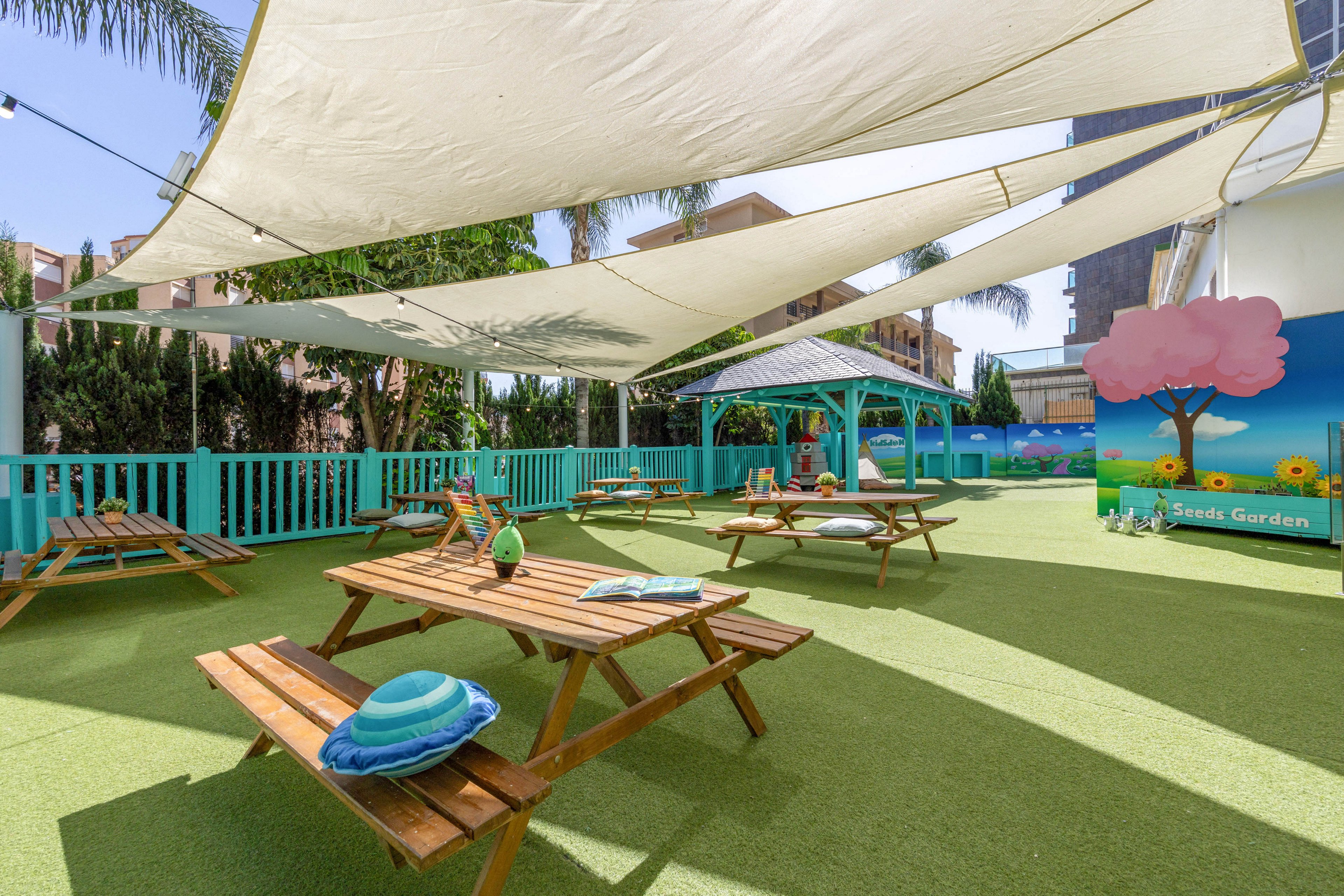 An outdoor play area features wooden picnic tables arranged on bright green artificial grass. Large white shade sails stretch overhead, creating a cool, inviting space. A turquoise fence and gazebo frame the area, with colorful cartoon wall art and toys adding a playful touch. The setting appears calm and ready for children’s activities in a sunny urban environment.