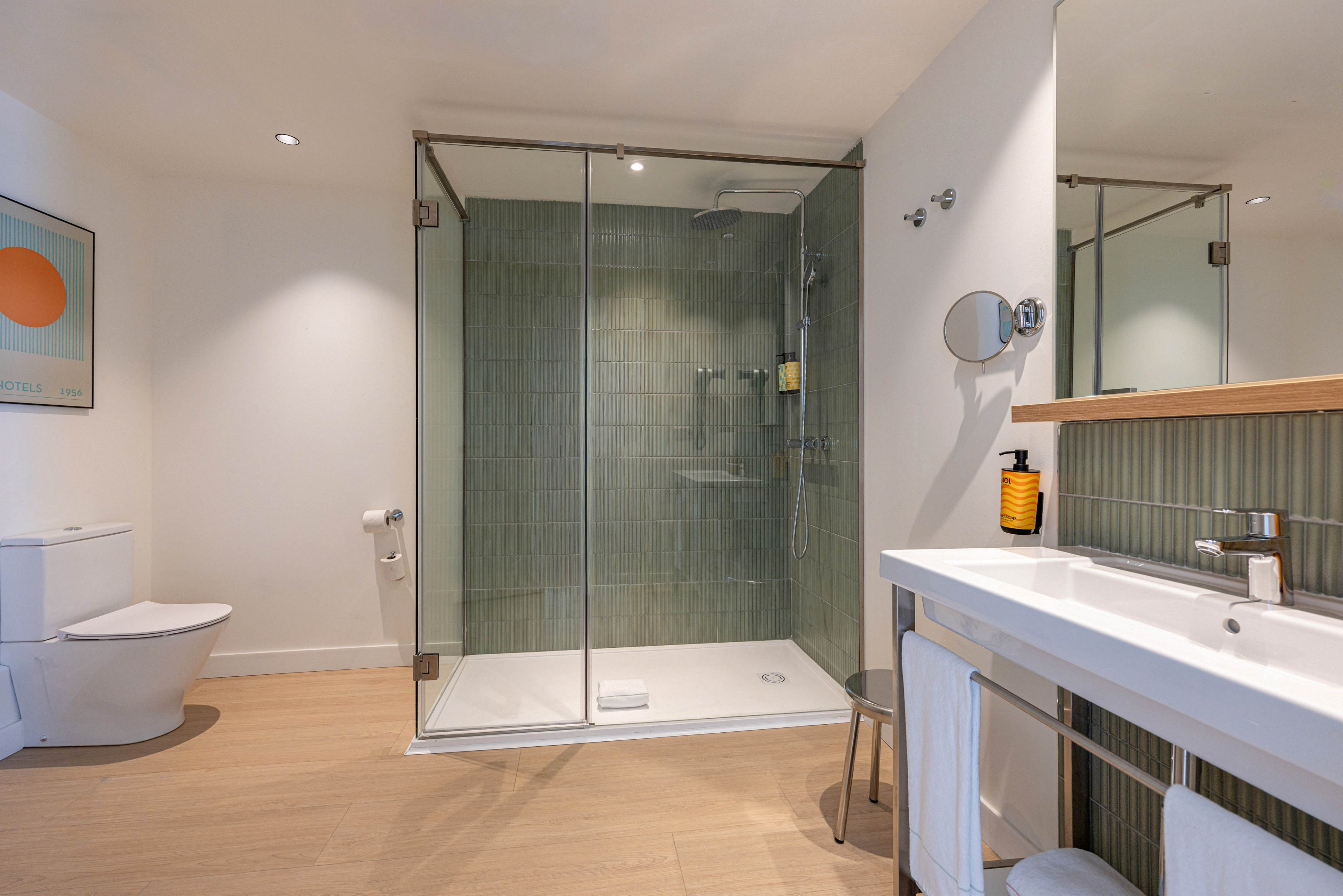 Contemporary bathroom interior featuring a large glass-enclosed walk-in shower with green tiled walls. A sleek white vanity with integrated sink and chrome faucet stands to the right, accessorized with a soap dispenser and towels. On the left, a modern toilet and wall-mounted toilet paper holder sit against a clean white wall. Warm wood flooring and recessed ceiling lights create a bright, inviting atmosphere, complemented by a colorful abstract artwork.