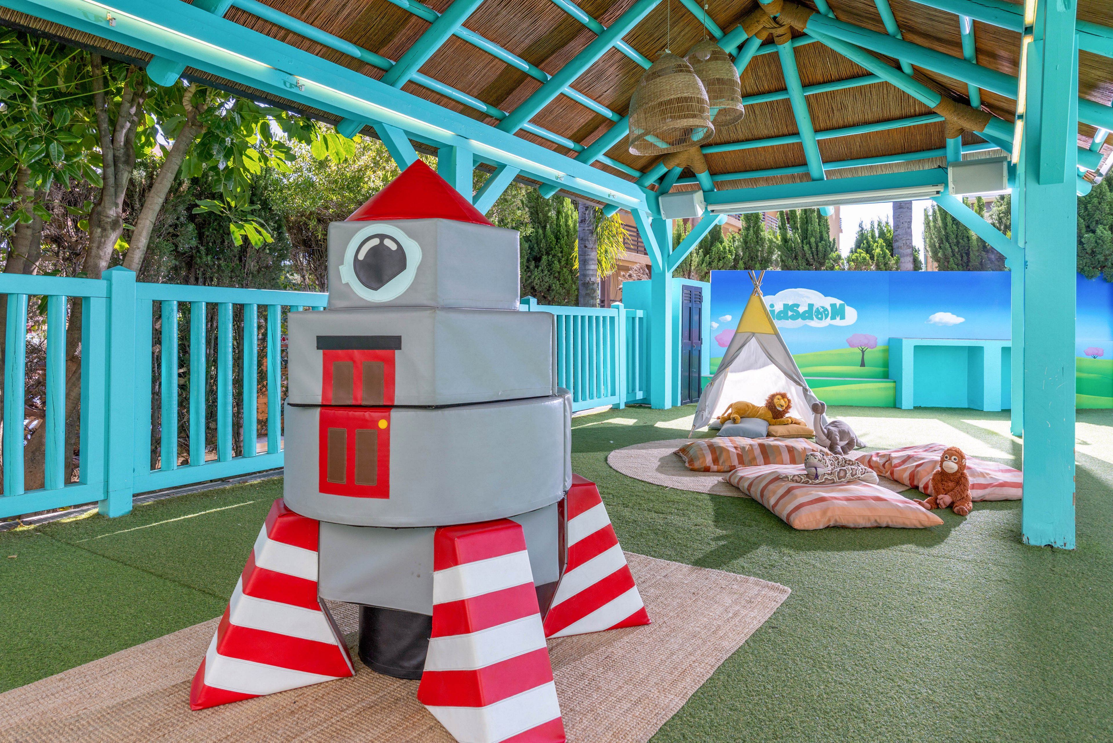 A brightly colored outdoor play area features a large soft rocket ship structure as the main focal point. The space is covered by a turquoise wooden pavilion with a thatched roof and surrounded by matching railings. In the background, a small teepee tent with cushions and stuffed animal toys creates a cozy corner for children. Artificial grass flooring and playful decor give the scene a cheerful, imaginative atmosphere.