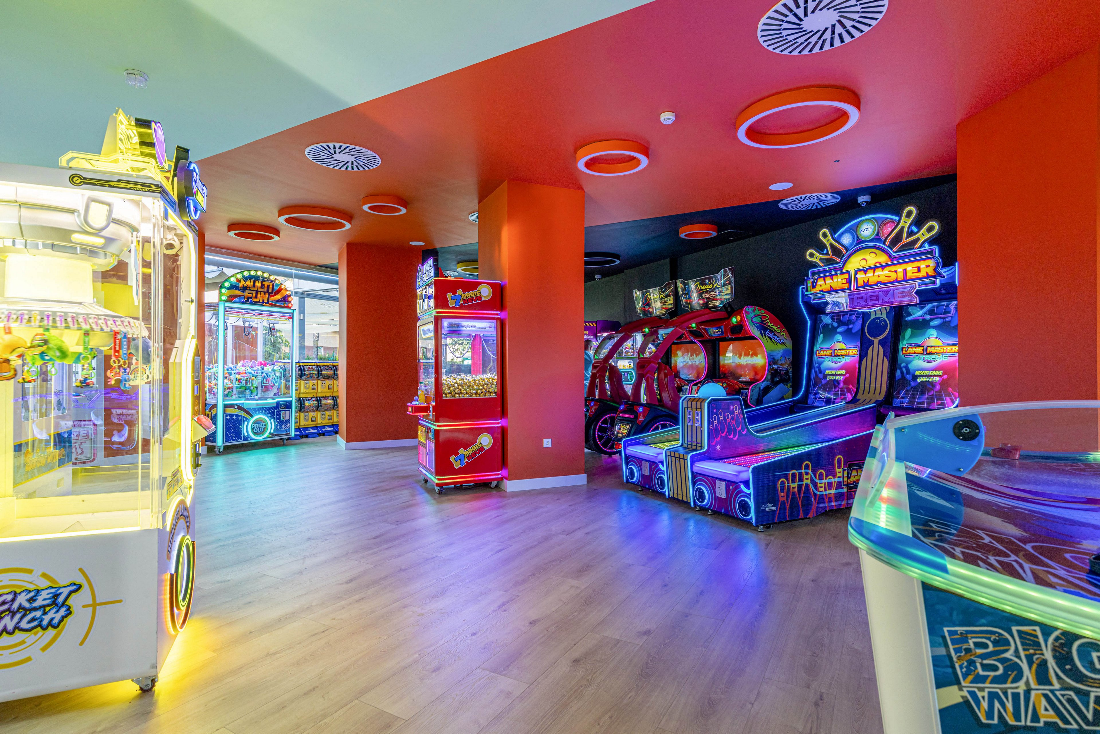 Brightly lit arcade interior featuring multiple colorful game machines. The space has red walls, a light wooden floor, and a ceiling with circular recessed fixtures. Various illuminated arcade cabinets and racing games line the back wall, creating a vibrant, playful atmosphere. The scene appears empty of people, emphasizing the machines and neon lights.