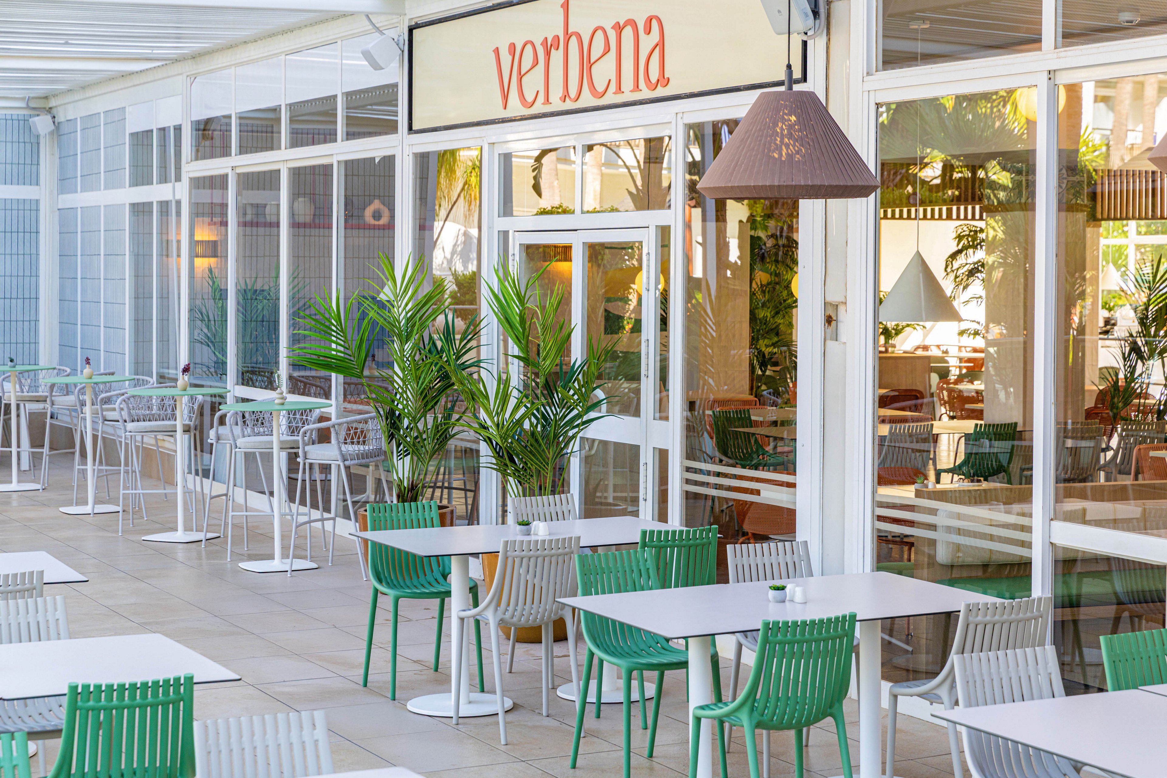 Bright outdoor seating area of a cafe named verbena features rows of white tables with green and gray chairs. Large glass windows reveal the interior space and reflect surrounding light. Potted palm plants add greenery and a relaxed, contemporary atmosphere. The terrace appears clean, airy, and ready to welcome guests.