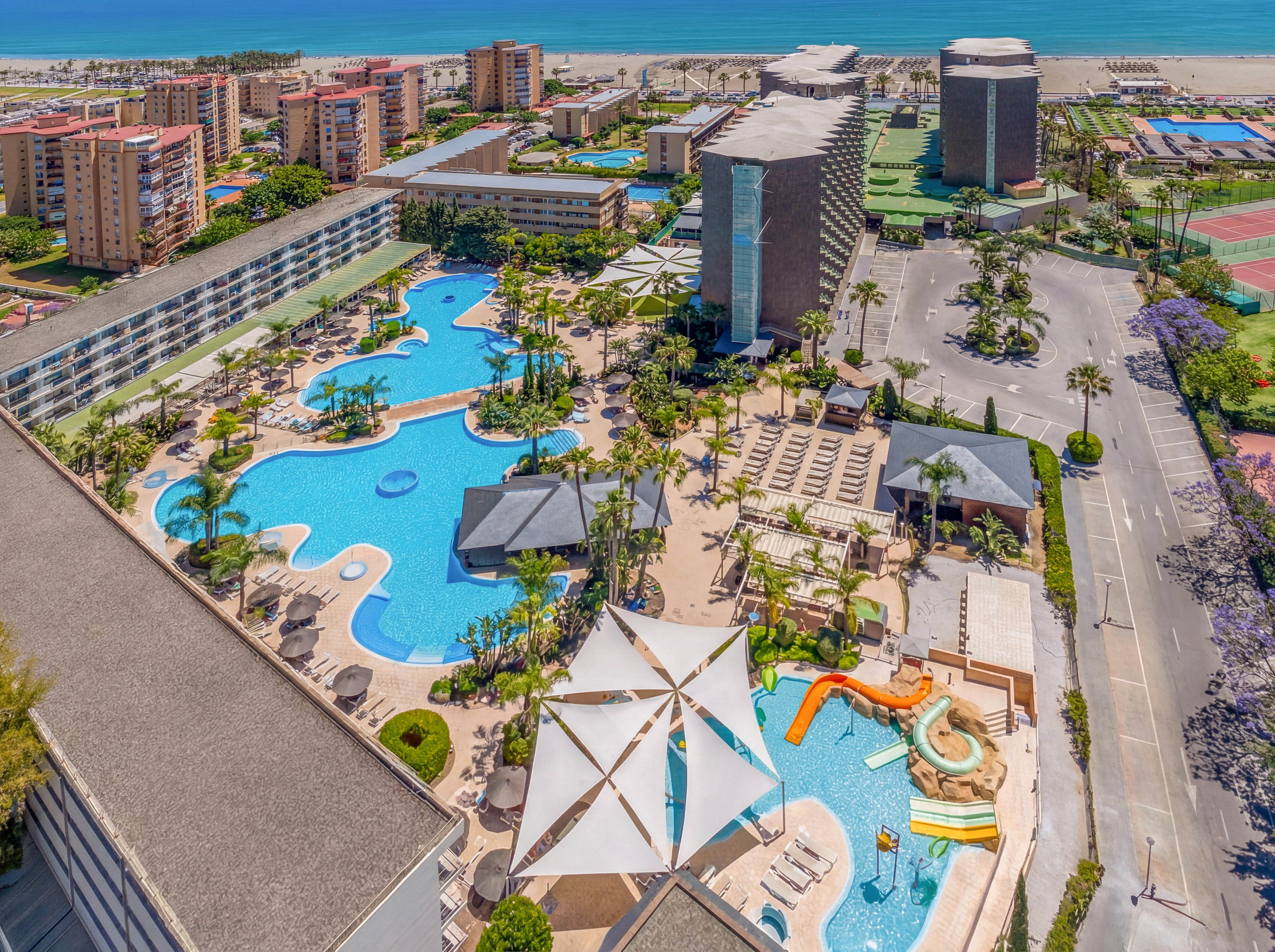 A large resort complex features an expansive, lagoon-style swimming pool with winding shapes and bridges. Several water slides and shallow play areas are visible, along with sun loungers neatly arranged around the pool. Tall hotel buildings and landscaped gardens surround the pool area, extending toward tennis courts and other recreational spaces. In the distance, a sandy beach and the ocean create a scenic coastal backdrop under clear skies.