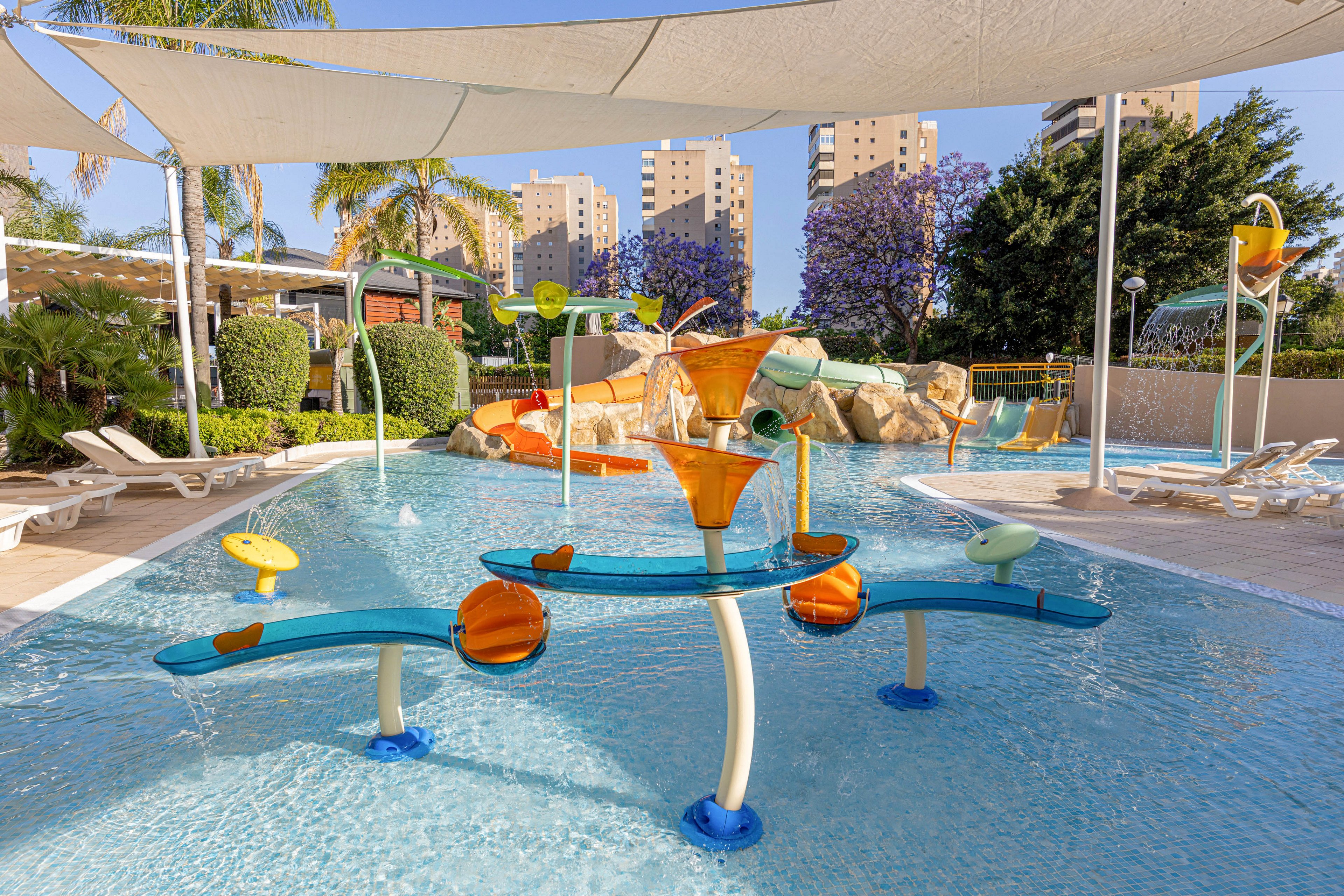 Colorful water play structures stand in a shallow outdoor splash pad pool. Tipping buckets and playful fountains are arranged under a large shade canopy. Lounge chairs and landscaped greenery surround the pool area, with tall residential buildings visible in the background. The bright daylight and clear water create a cheerful, family-friendly atmosphere.