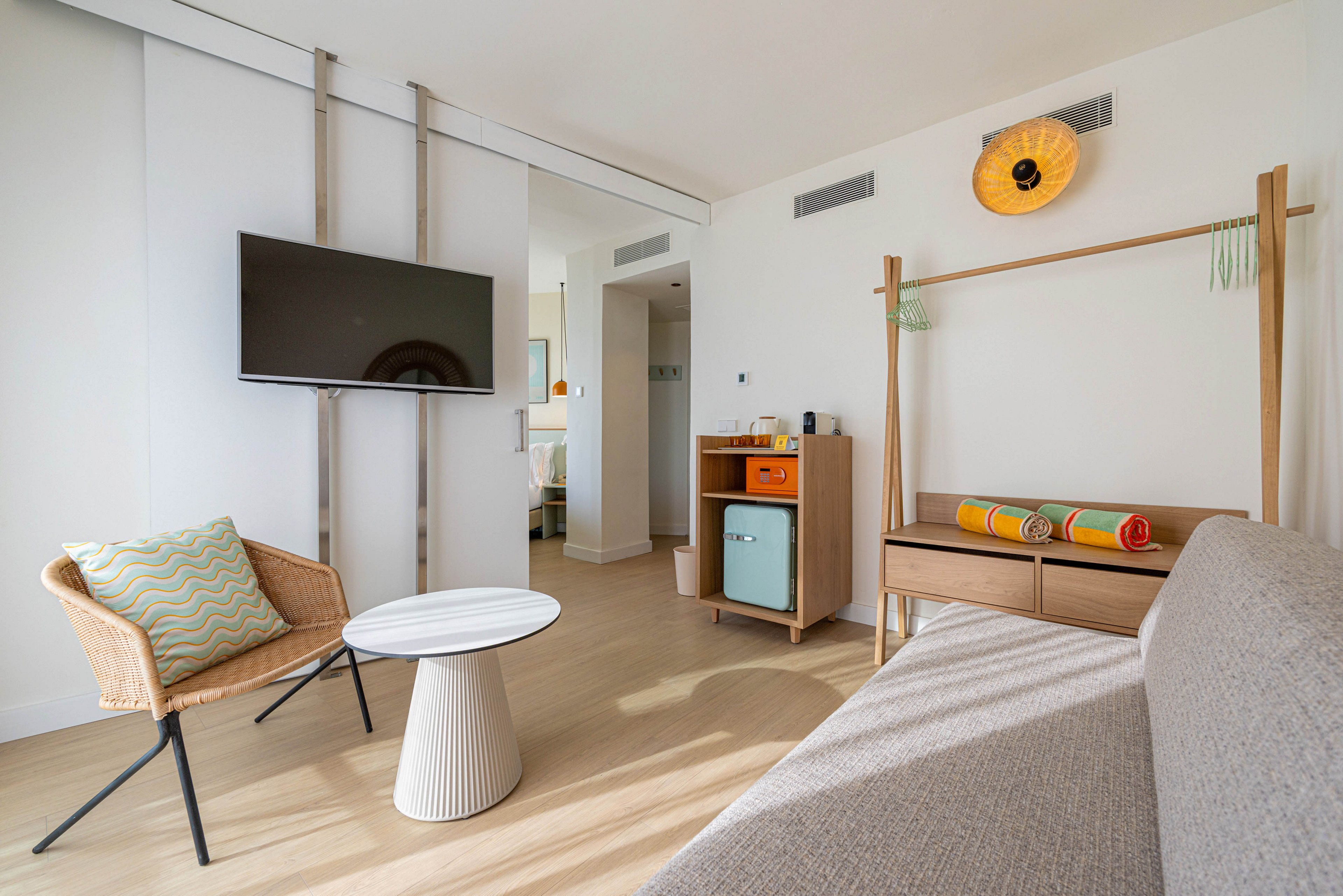 A bright modern hotel room features a wall-mounted flat-screen TV opposite a light gray sofa. A woven chair with a chevron cushion and a small round side table sit on a light wood floor. In the background, a compact cabinet with a pastel-colored mini fridge and open shelving adds a retro accent. Rolled towels and a decorative wall light contribute to the clean, minimalist aesthetic of the space.
