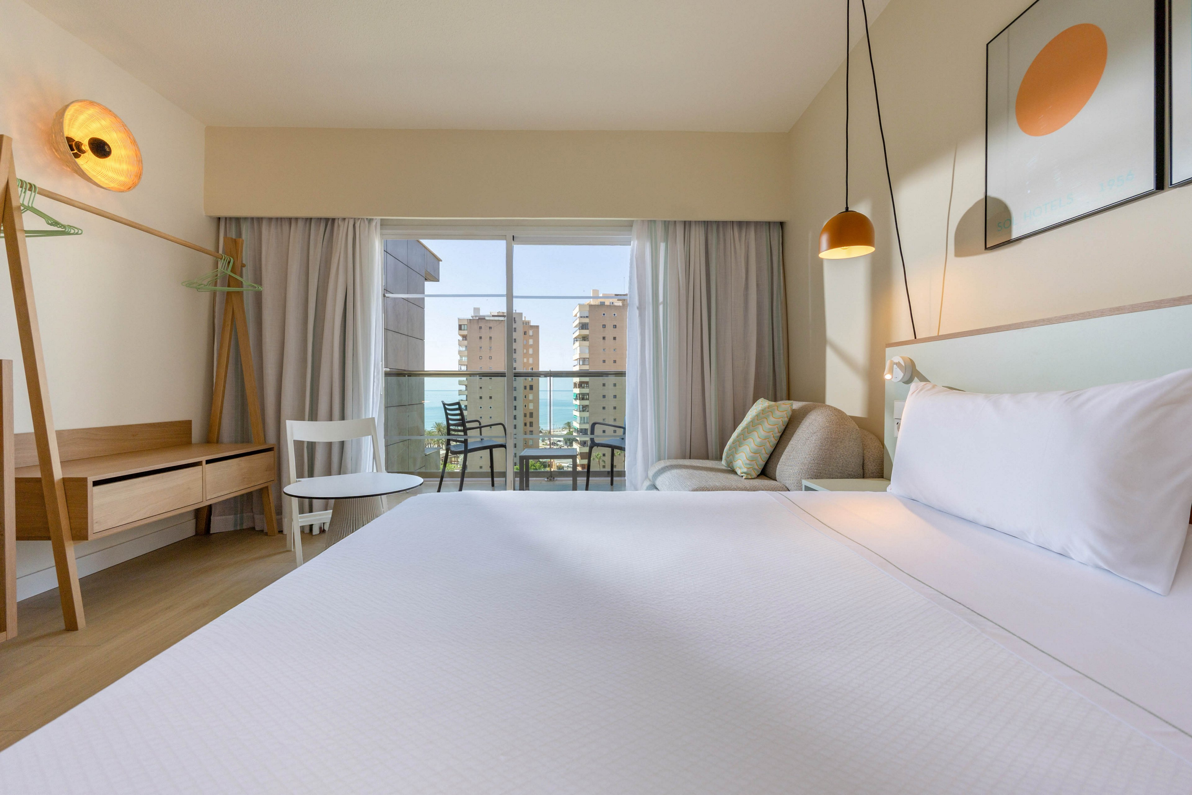 A bright modern hotel room features a large neatly made bed in the foreground. Minimalist wooden furniture, including a desk, chair, and open wardrobe, lines the left side of the room. Soft pendant lighting and abstract wall art add a contemporary touch to the neutral decor. A glass door opens to a balcony with two chairs and a table, overlooking tall residential buildings in the distance.