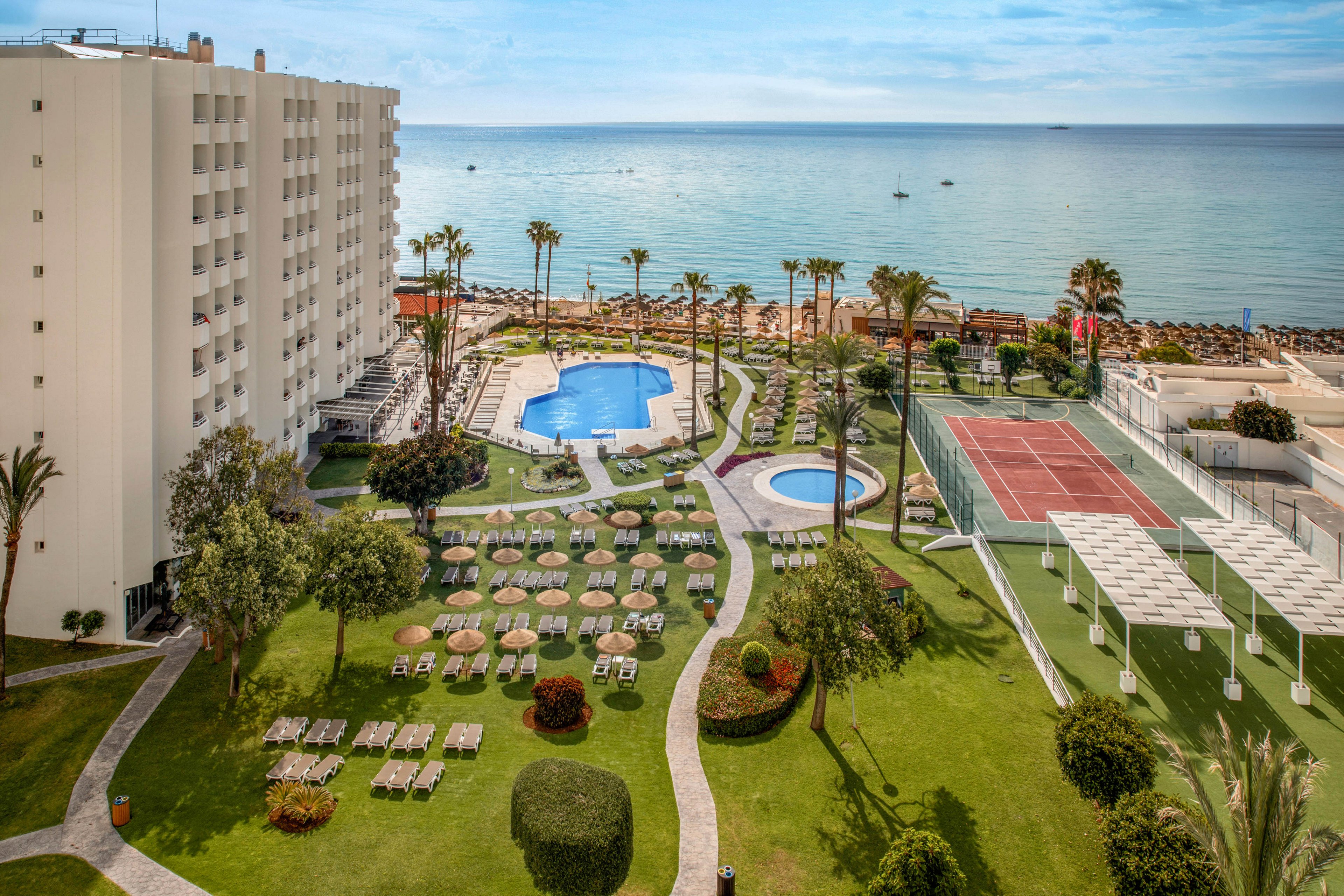 A coastal resort overlooks a calm sea with a sandy beach and sun loungers. In the foreground, a landscaped garden features rows of deck chairs, palm trees, and manicured lawns. A large outdoor swimming pool and a smaller circular pool sit near the beach, surrounded by terraces and pathways. To the right, red tennis courts and shaded structures provide additional recreational space beside a multi-story hotel building.