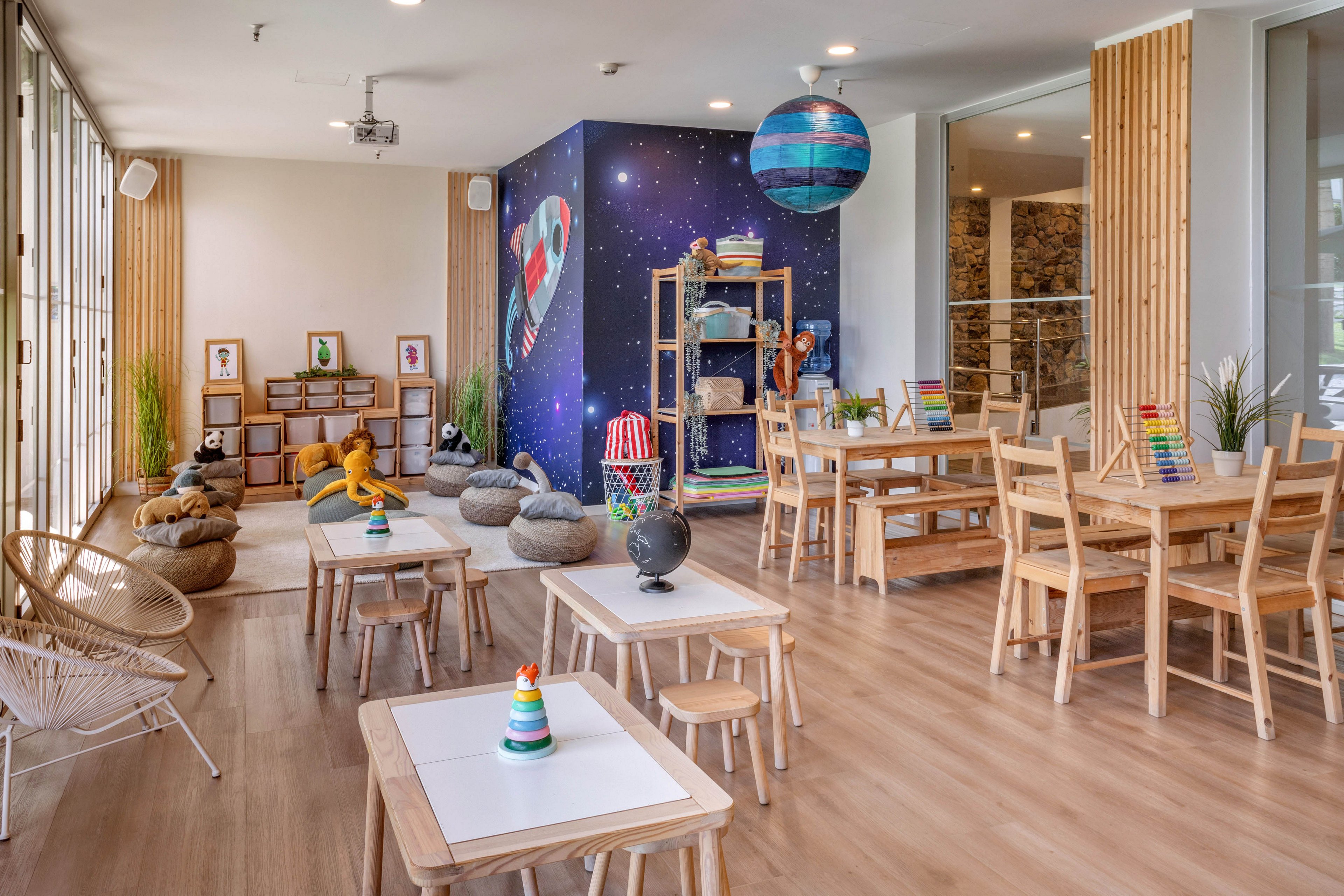 Bright, modern children's playroom featuring wooden tables, chairs, and educational toys. A colorful space-themed mural with planets and a rocket decorates the back wall, creating a playful focal point. Soft seating cushions and beanbags are arranged in a cozy reading or relaxation corner. Large windows bring in natural light, highlighting the clean, minimalist design and warm wood flooring.