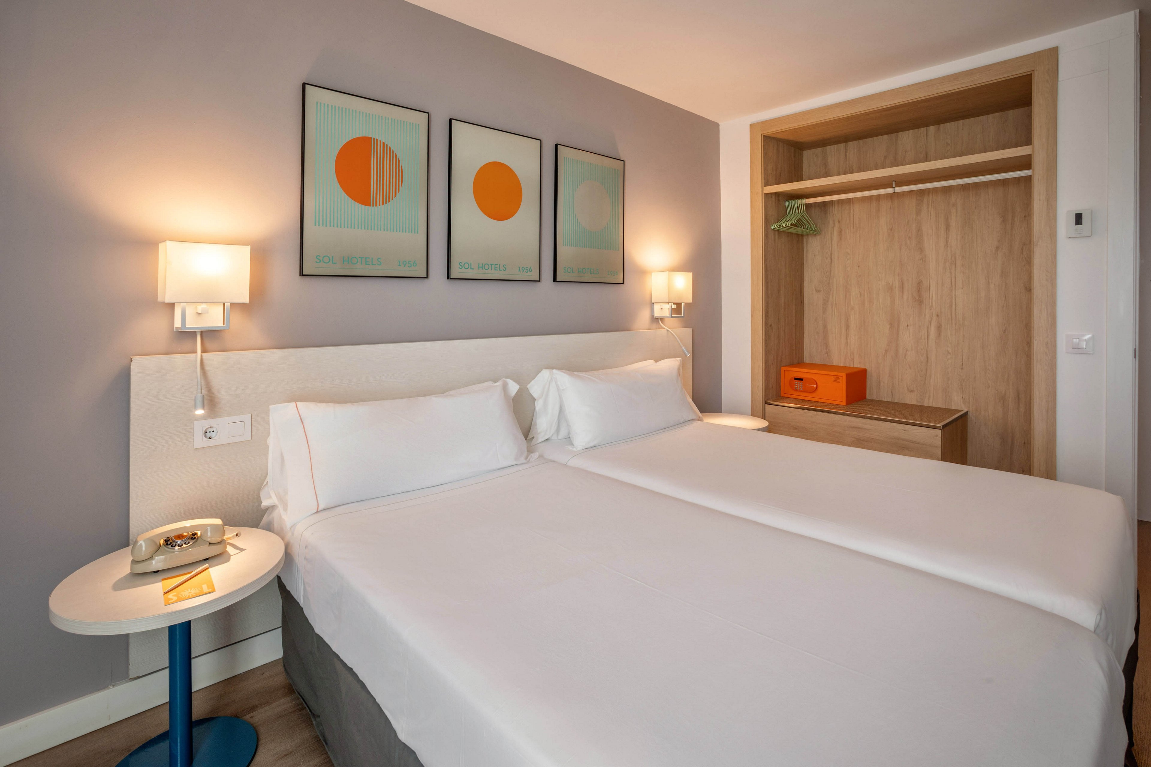 Contemporary hotel room featuring two neatly made twin beds with white linens against a soft gray wall. The space includes wall-mounted bedside lamps, a small round side table, and an open wooden wardrobe with hangers and a storage box. Three minimalist art prints with circular designs add color above the headboard. The room appears clean, bright, and inviting, suggesting a comfortable stay for travelers.