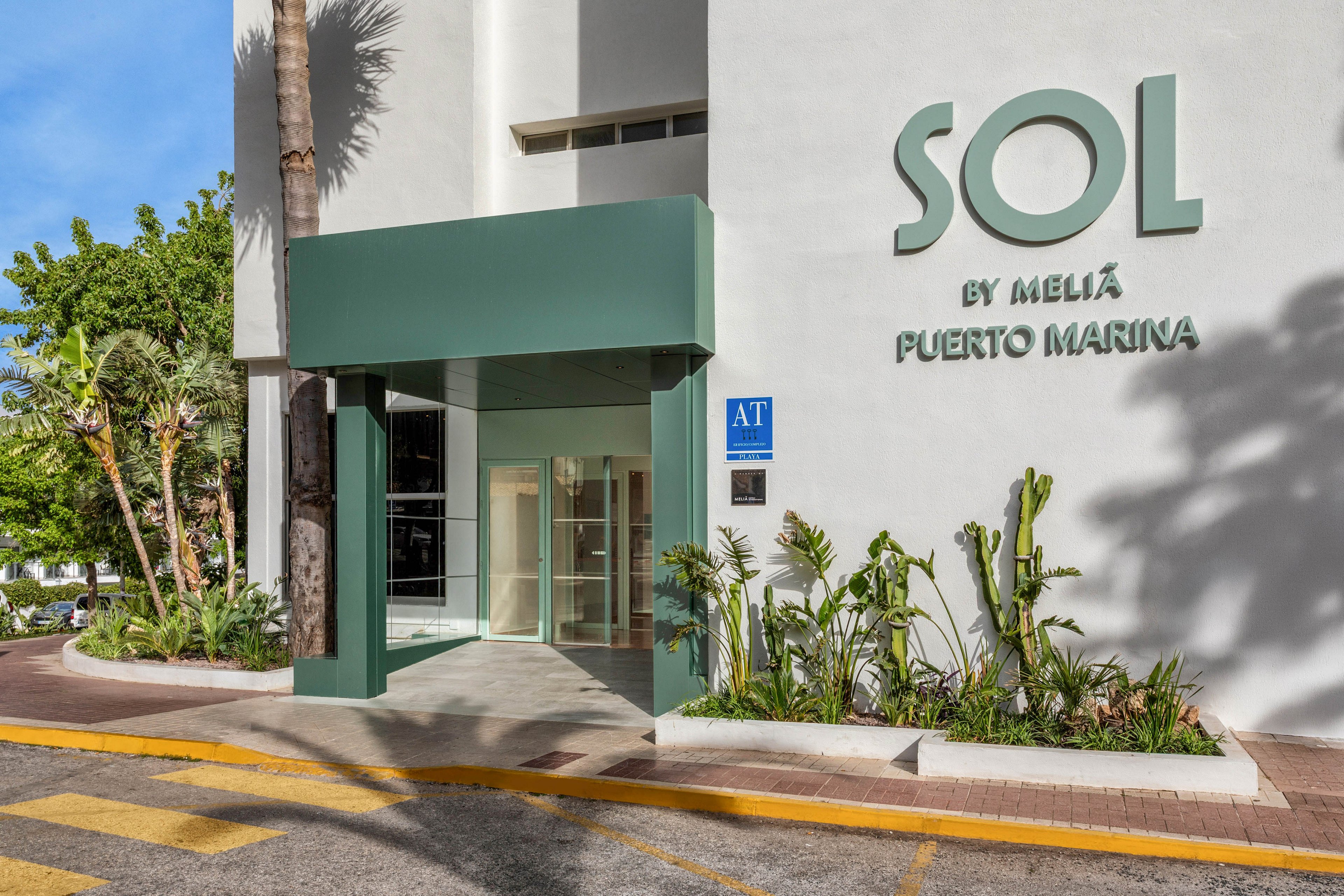 Modern entrance of the Sol by Meliá Puerto Marina hotel with a green canopy and glass doors. The building facade is white with the hotel name clearly displayed on the wall. Tropical plants and palm trees frame the walkway and add greenery to the scene. A quiet street with yellow road markings runs in front of the hotel, suggesting an urban coastal setting.