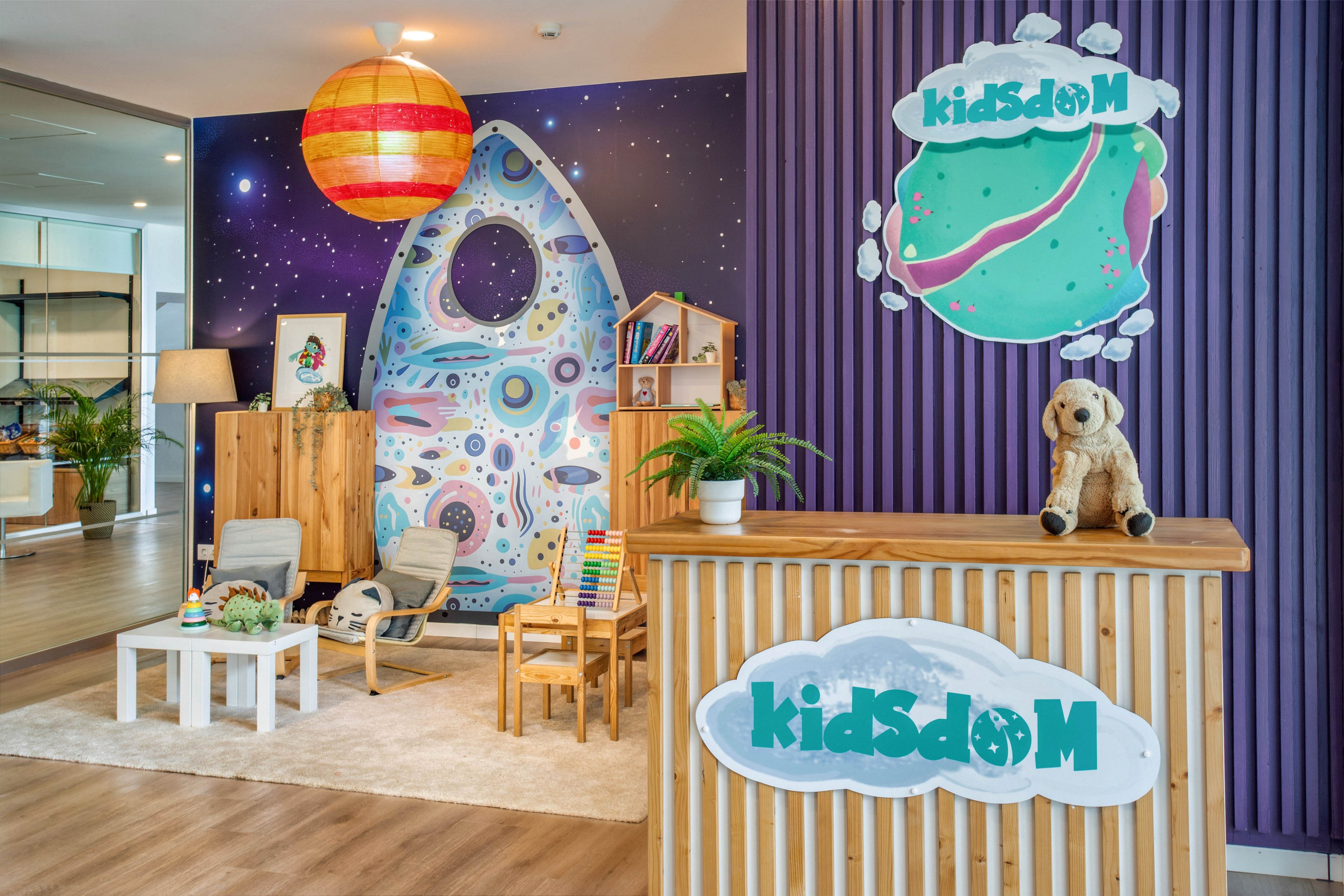 A bright children’s play area features a wooden reception counter with a plush dog toy and a sign reading “kidsdom.” Behind it, a large wall mural shows a whimsical planet and a rocket-shaped doorway set against a starry space background. A cozy seating corner with small chairs, a table, and bookshelves sits on a light rug, complemented by indoor plants. Warm wooden flooring and a large hanging lamp create a welcoming, playful atmosphere.