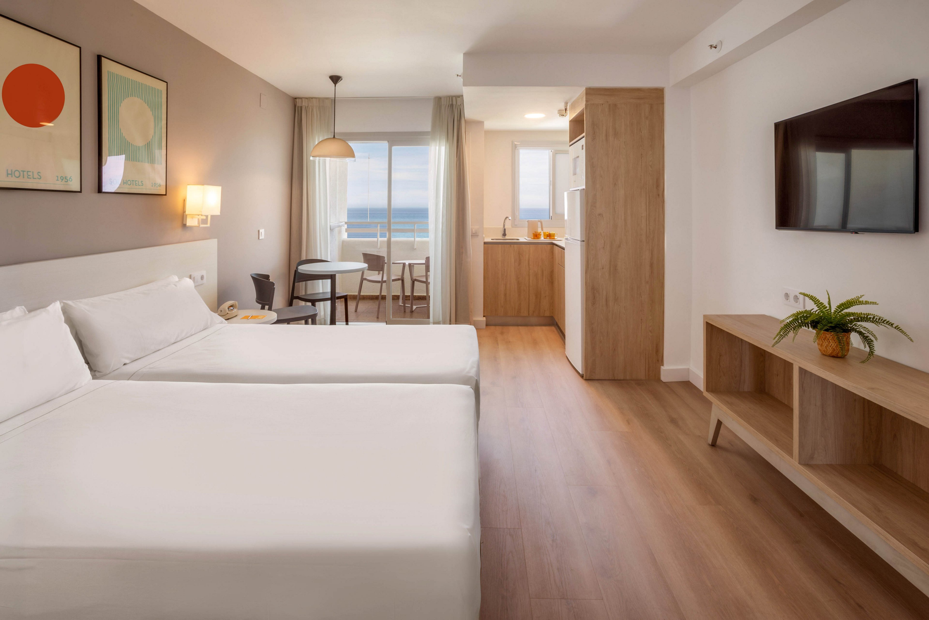 Contemporary hotel room interior featuring two neatly made twin beds with white linens. The space includes a small kitchenette, wall-mounted television, and wooden furniture with clean lines. A dining table and chairs sit near large glass doors that open to a balcony overlooking the ocean. Soft neutral tones, minimalist artwork, and natural light create a calm, inviting atmosphere.