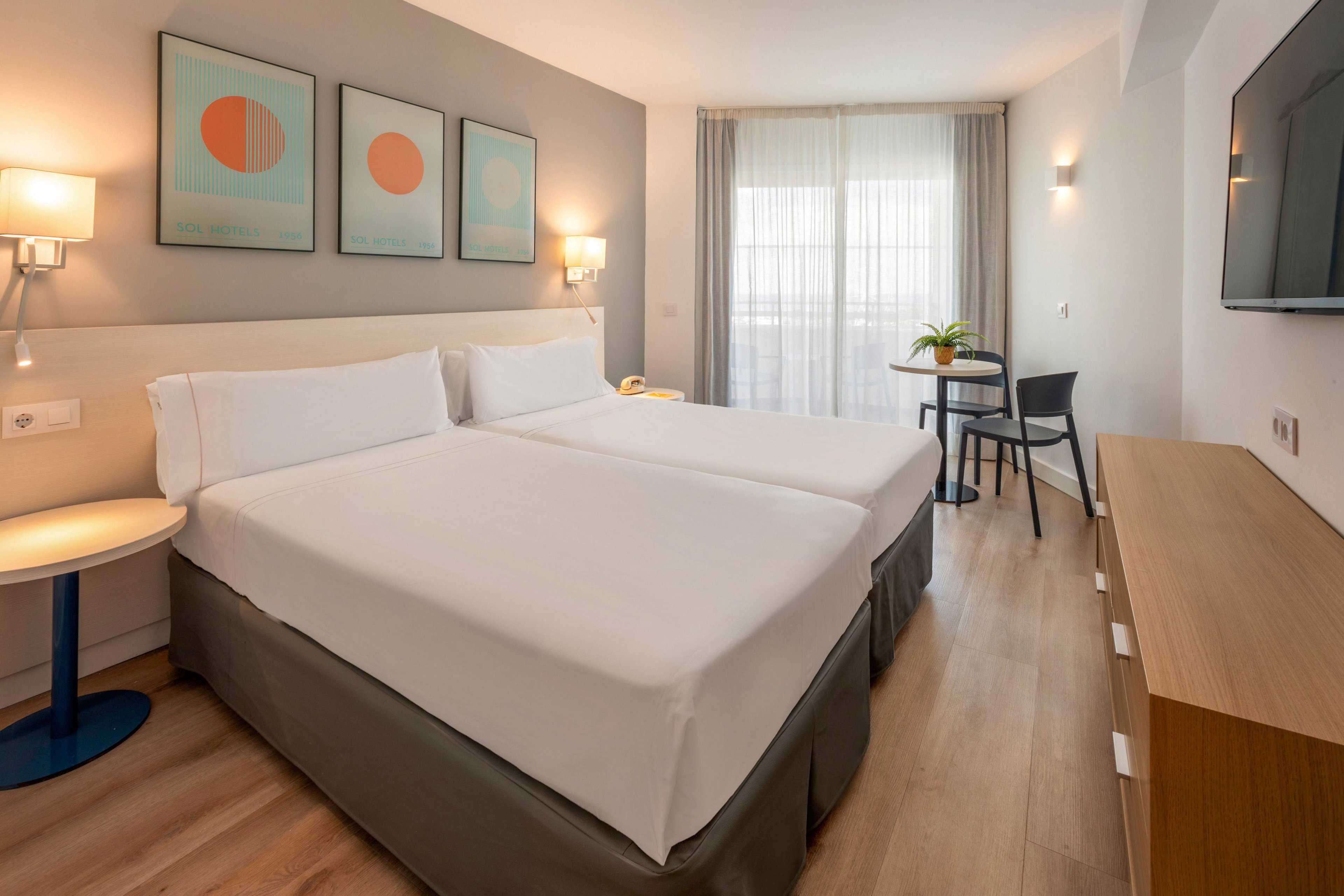 A contemporary hotel room features two neatly made twin beds with white linens and a gray bed base. The space includes light wood flooring, a wall-mounted flat-screen TV, and minimalist bedside lighting. Three abstract art prints hang above the headboard, while a small round table and chair sit near the window with sheer curtains. Overall, the room feels bright, clean, and inviting with a neutral color palette.