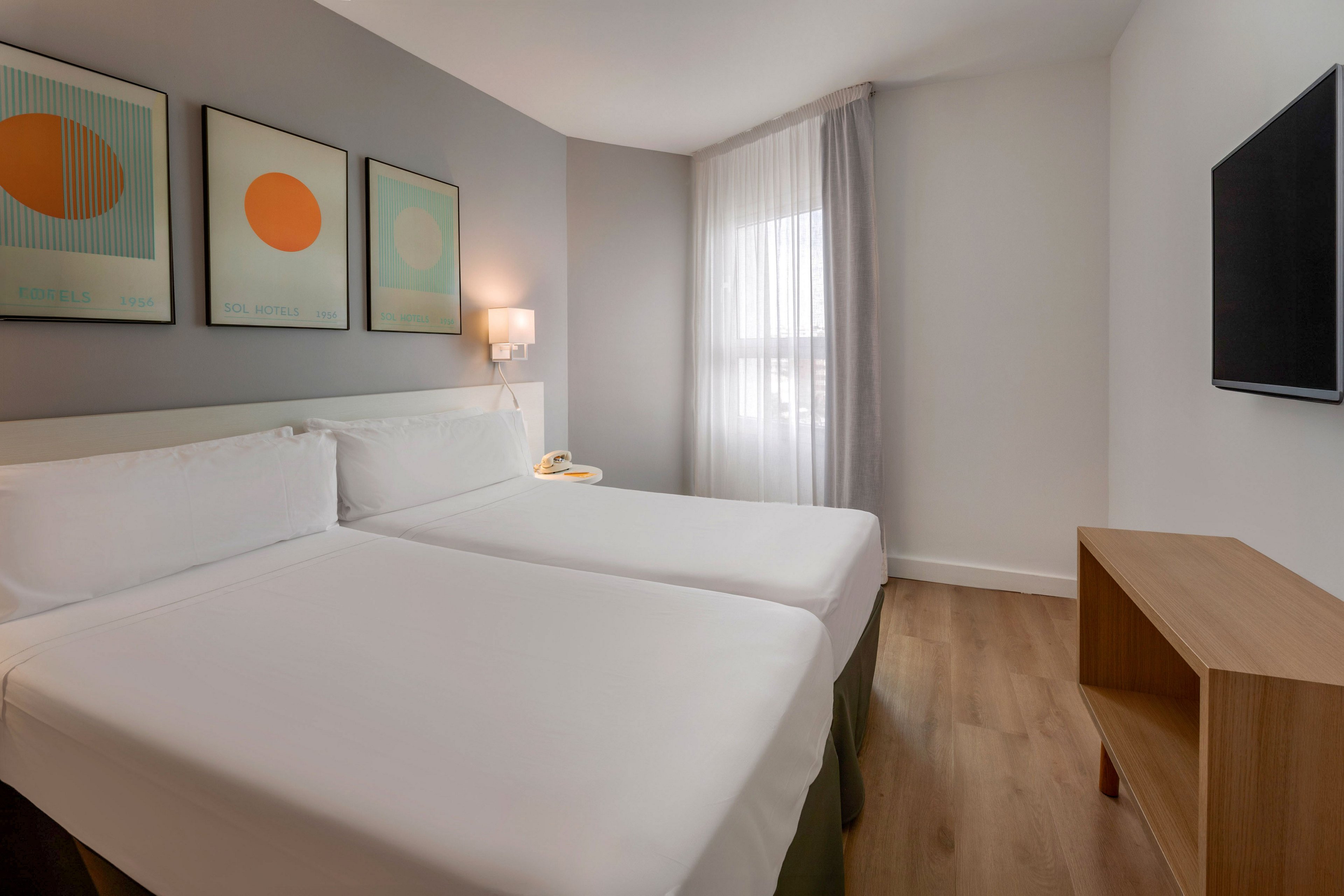 A contemporary hotel room features two neatly made twin beds with white linens. The space is decorated with three abstract art prints above the headboard and a wall-mounted lamp between the beds. Natural light filters through sheer curtains covering a large window, brightening the neutral-toned walls and wooden floor. A flat-screen television is mounted on the wall opposite the beds, above a simple wooden console.