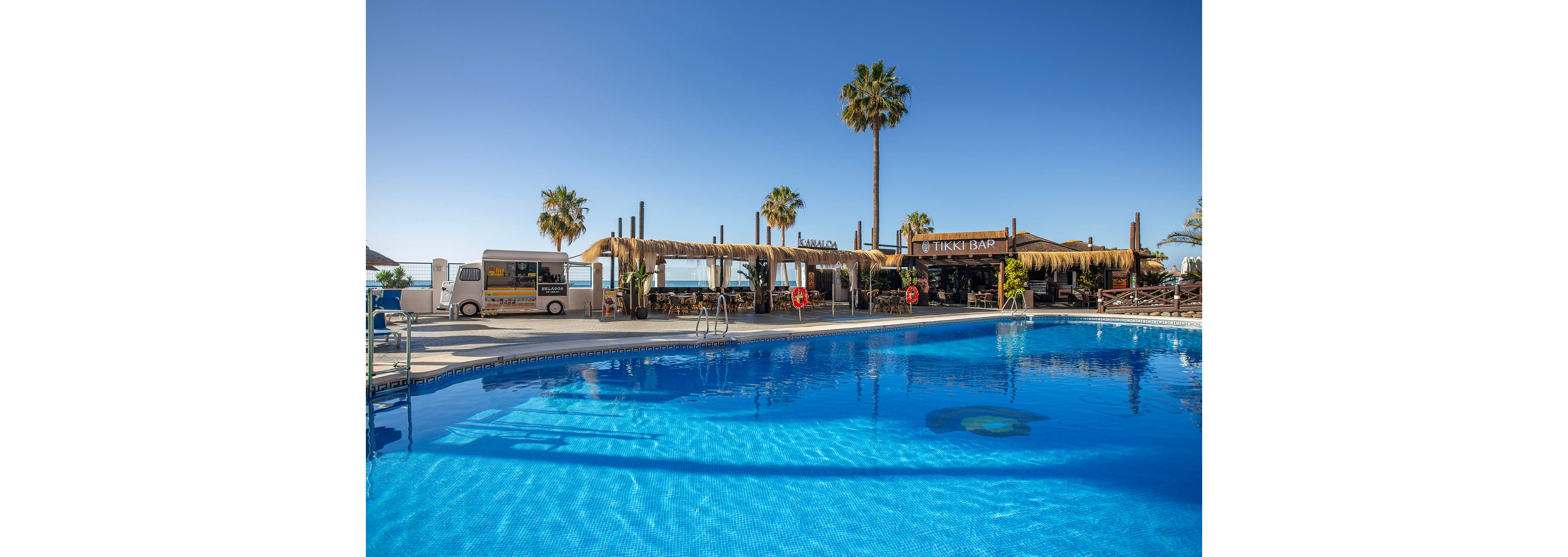 A vibrant outdoor pool is set in front of a tiki bar with thatched roofing, surrounded by tall palm trees under a clear blue sky. The scene evokes a relaxed, tropical atmosphere, with visible signage reading 'TIKKI BAR' and a white van parked nearby. The water in the pool is bright blue, reflecting the sunny weather, and red life preservers are mounted along the poolside. The setting suggests a leisure or vacation destination, ideal for relaxation and social gatherings.