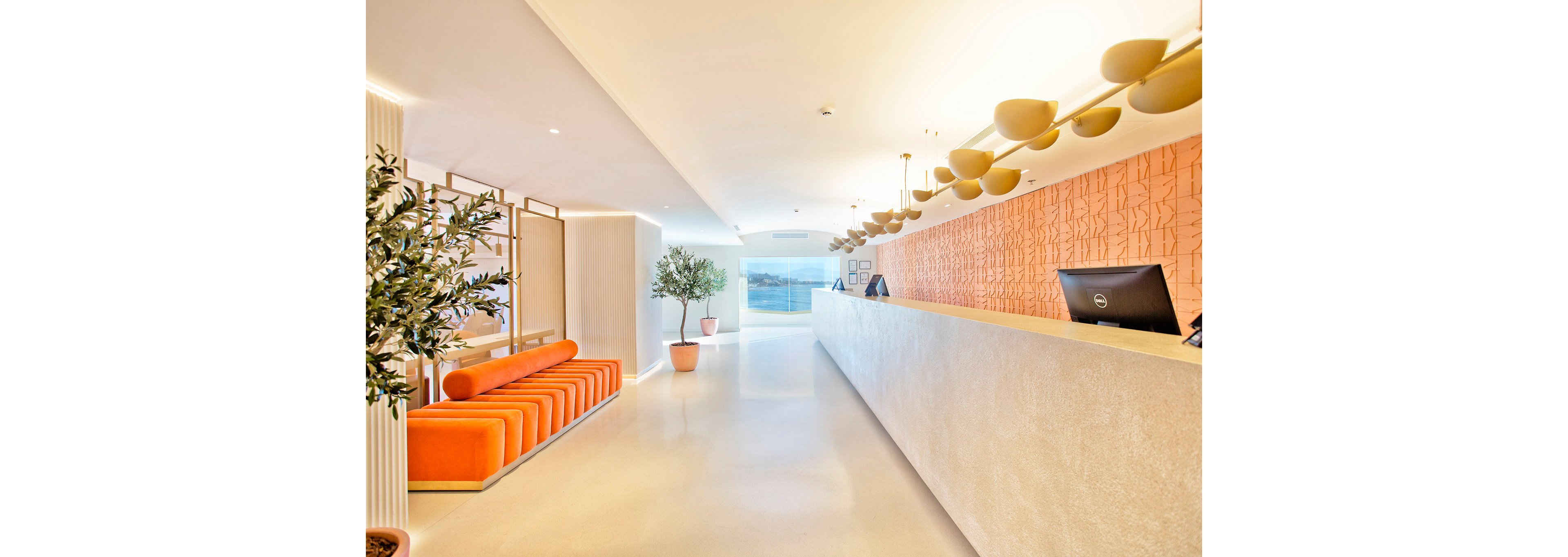 A bright, contemporary hotel reception area features a long white counter and a row of orange cushioned benches. The space is accented with potted plants and a textured orange wall behind the desk. Pendant lights hang from the ceiling, and a computer monitor is visible on the counter. Natural light floods in from large windows at the far end, creating an inviting atmosphere.