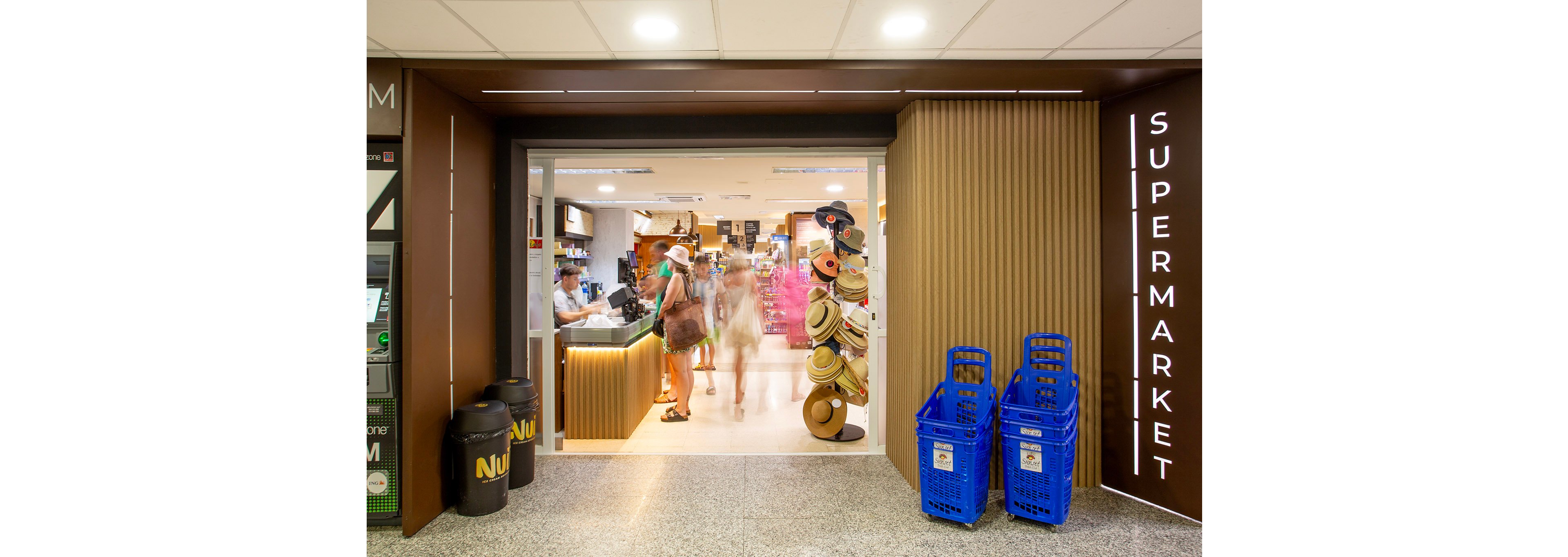 A contemporary supermarket entrance features two blue plastic shopping baskets and a display of hats on a stand. Several people, including adults and children, are visible inside, some at the checkout counter. The setting is brightly lit with a clean, organized interior and the word 'SUPERMARKET' clearly visible on the wall. Two black bins with the text 'NUI' are placed near the entrance. The overall mood is busy and welcoming.