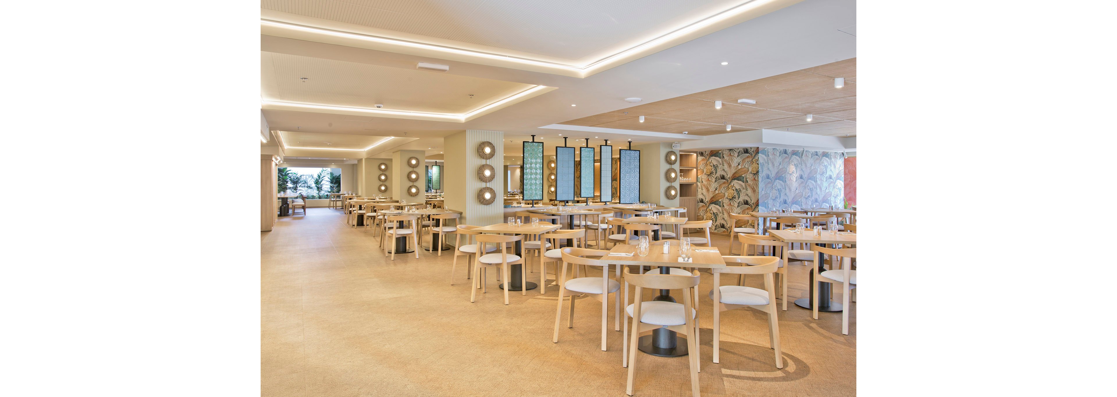 Spacious restaurant dining area featuring light wood tables and chairs arranged neatly. The setting is bright with natural and artificial lighting, complemented by decorative wall art and mirrors. The overall mood is calm and inviting, with a neutral color palette and clean lines. No people are present, emphasizing the serene and organized atmosphere.