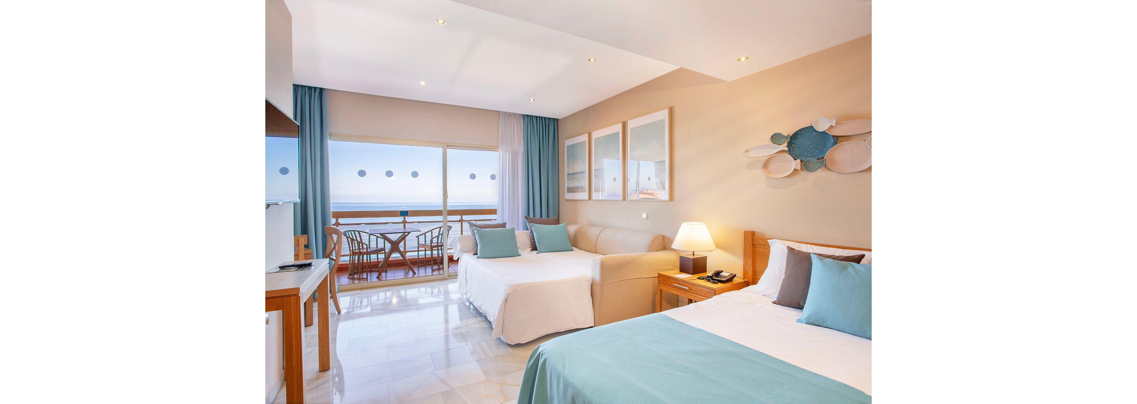 A spacious hotel room features two beds, a sofa, and a balcony overlooking the ocean. The setting is airy and inviting, with soft blue and beige tones, contemporary decor, and natural light streaming in. The room includes a small table with chairs on the balcony, framed wall art, and a bedside lamp. The overall mood is relaxed and vacation-ready, ideal for leisure travelers.