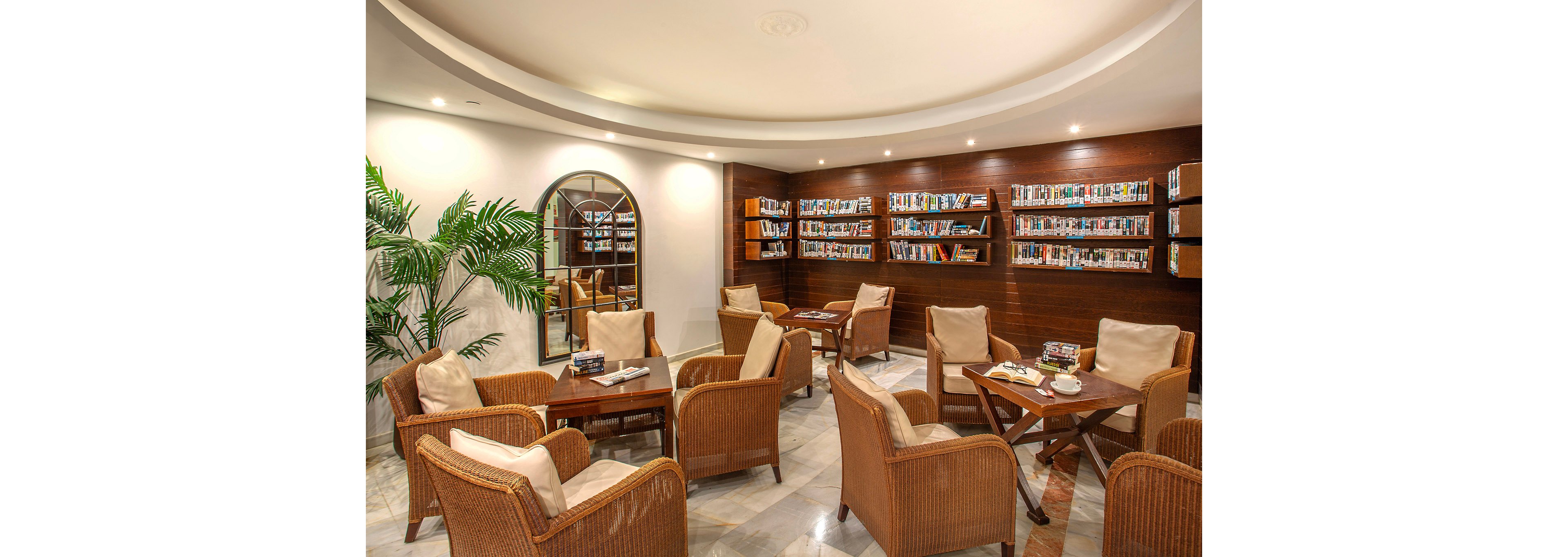 A warmly lit library lounge features wicker armchairs with beige cushions arranged around wooden tables. The setting includes wall-mounted bookshelves filled with books, a large decorative plant, and a round wall mirror. The marble floor and soft lighting create a relaxed, inviting atmosphere for reading or conversation. No visible text or numbers are present in the image.