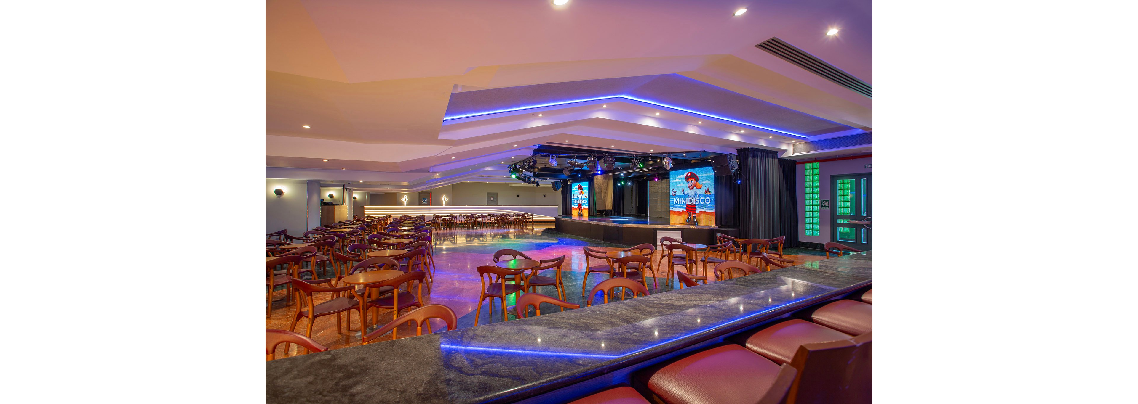 A spacious, contemporary event hall features a central stage with lighting and two large digital screens displaying 'MINIDISCO.' The room is filled with wooden chairs and tables, arranged for an audience, and illuminated by blue accent lighting. The setting is empty, highlighting the polished floors and modern ceiling design. The visible text 'MINIDISCO' appears on both screens.