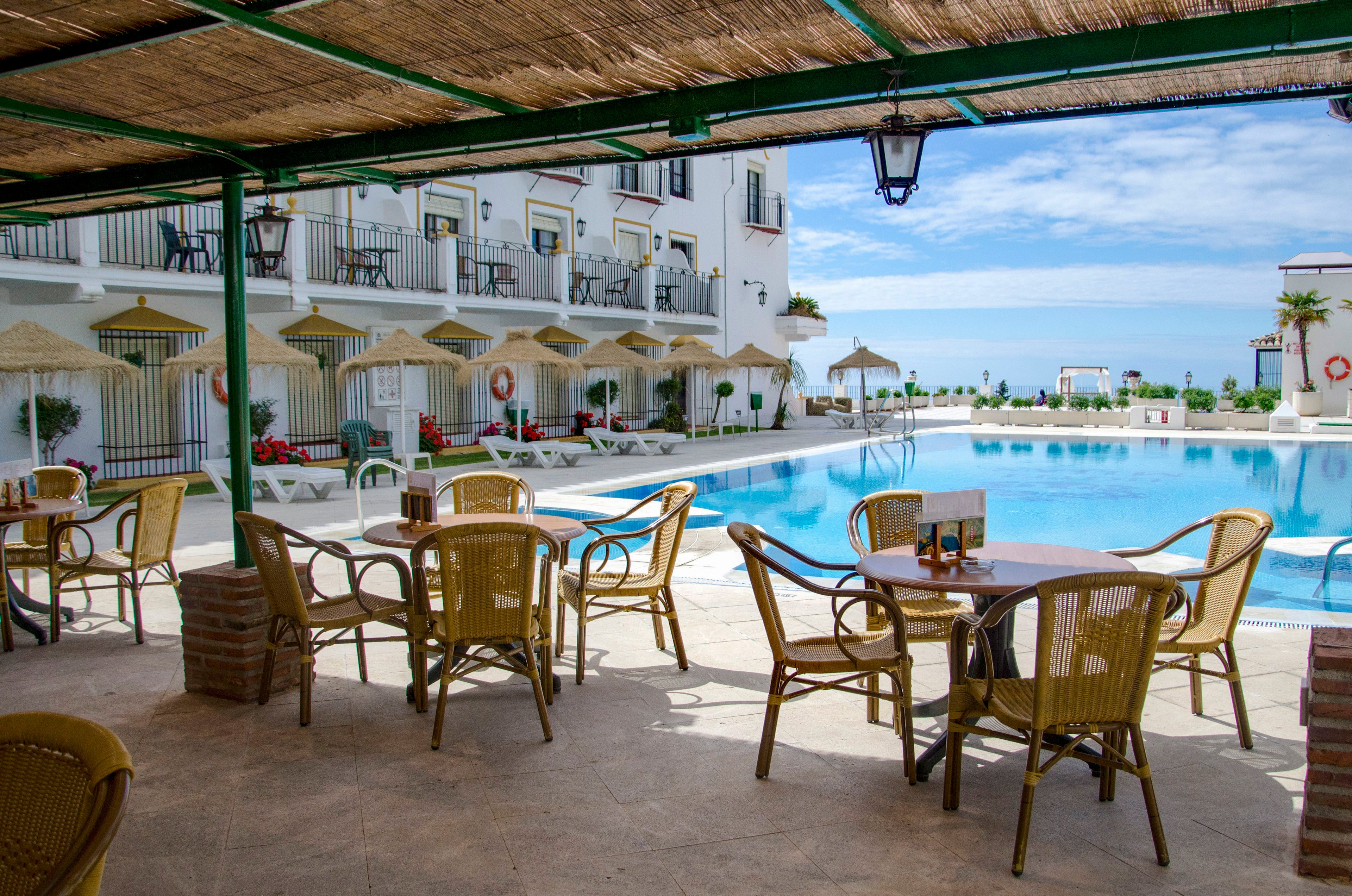 An open-air terrace with wicker chairs and tables faces a large outdoor swimming pool. White multi-story resort buildings with balconies and poolside umbrellas surround the water. The covered patio roof casts shade over the seating area, while bright sunlight illuminates the pool deck. In the distance, the blue sky and sea create a relaxing vacation atmosphere.