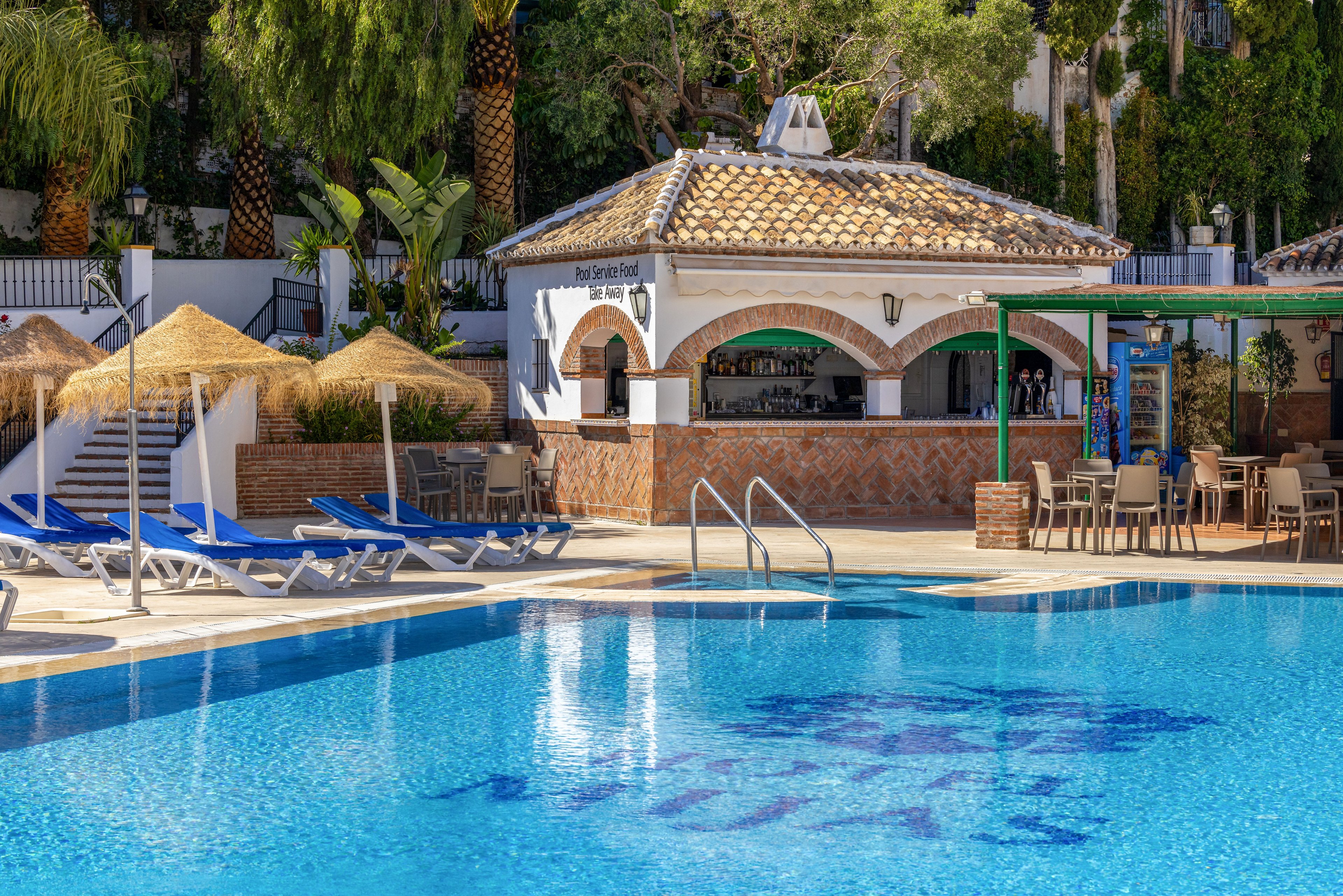 A clear blue swimming pool is shown in front of an outdoor bar with a tiled roof and arched openings. Blue lounge chairs line the poolside, accompanied by straw parasols and metal railings leading into the water. The bar area includes tables and chairs under a shaded structure, with lush green trees and plants in the background. Text is faintly visible on the pool floor, suggesting the name of the resort.