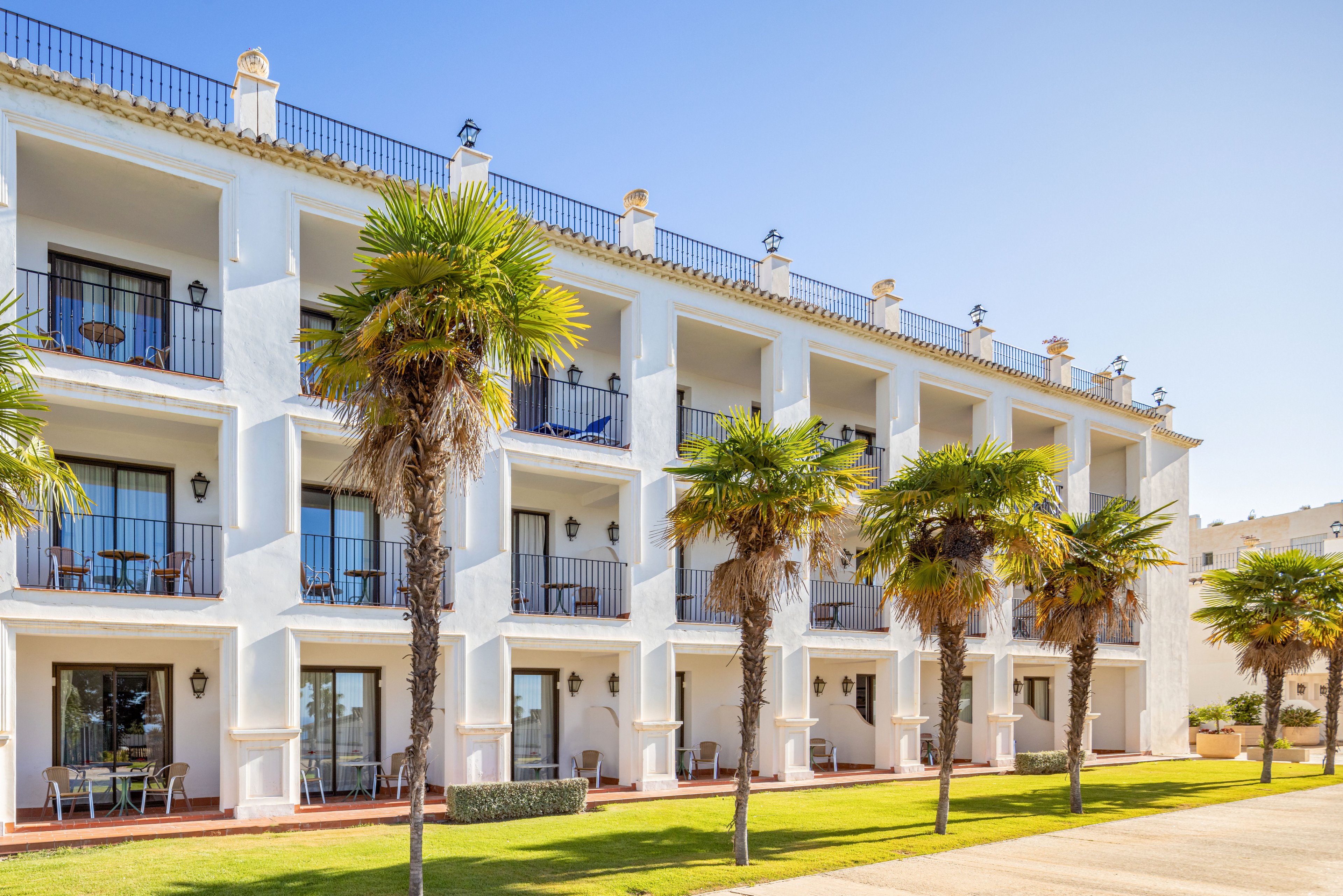 A white, multi-story hotel building features rows of balconies with outdoor seating. Tall palm trees line the front, adding a tropical touch to the sunny setting. The clear blue sky and bright light suggest a warm, coastal climate. The scene conveys a relaxed vacation atmosphere with clean architectural lines and open terraces.