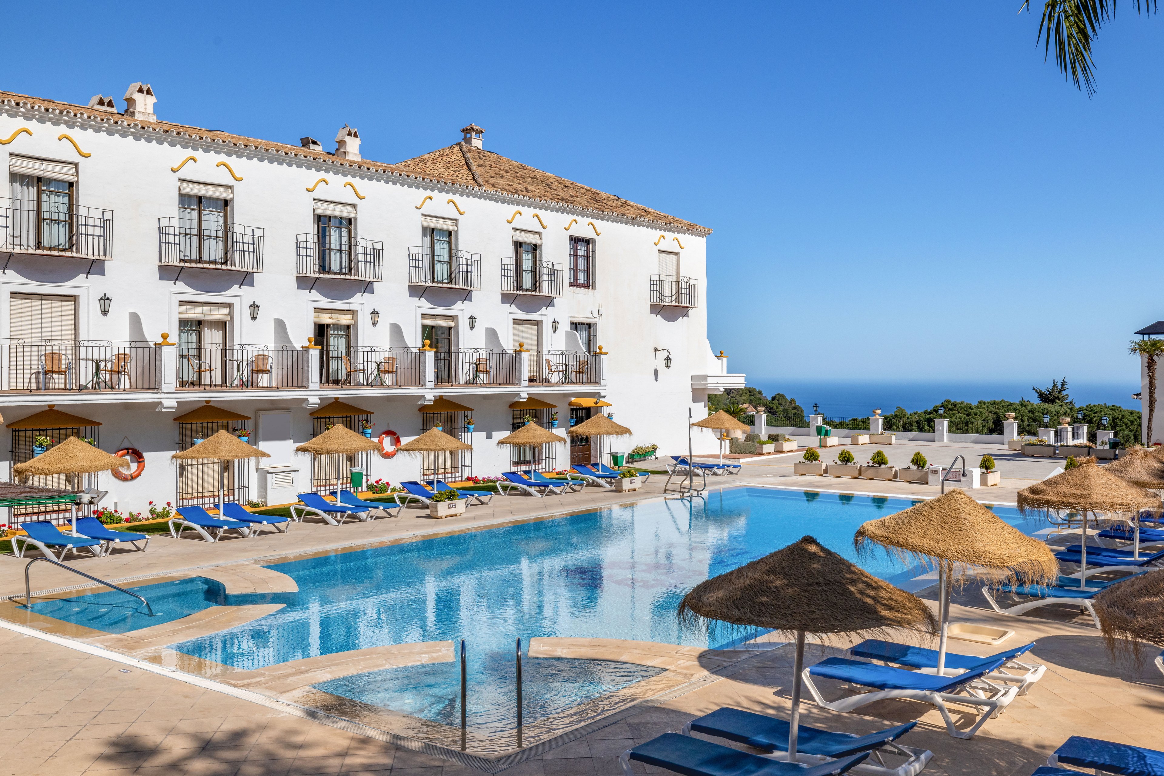A bright Mediterranean-style hotel overlooks a clear blue swimming pool surrounded by sun loungers and straw parasols. The scene is set on a sunny day with a vivid blue sky and distant ocean horizon. White stucco walls, wrought-iron balconies, and terracotta roof tiles create a classic coastal resort atmosphere. The tranquil setting suggests a relaxing vacation spot focused on leisure and outdoor enjoyment.