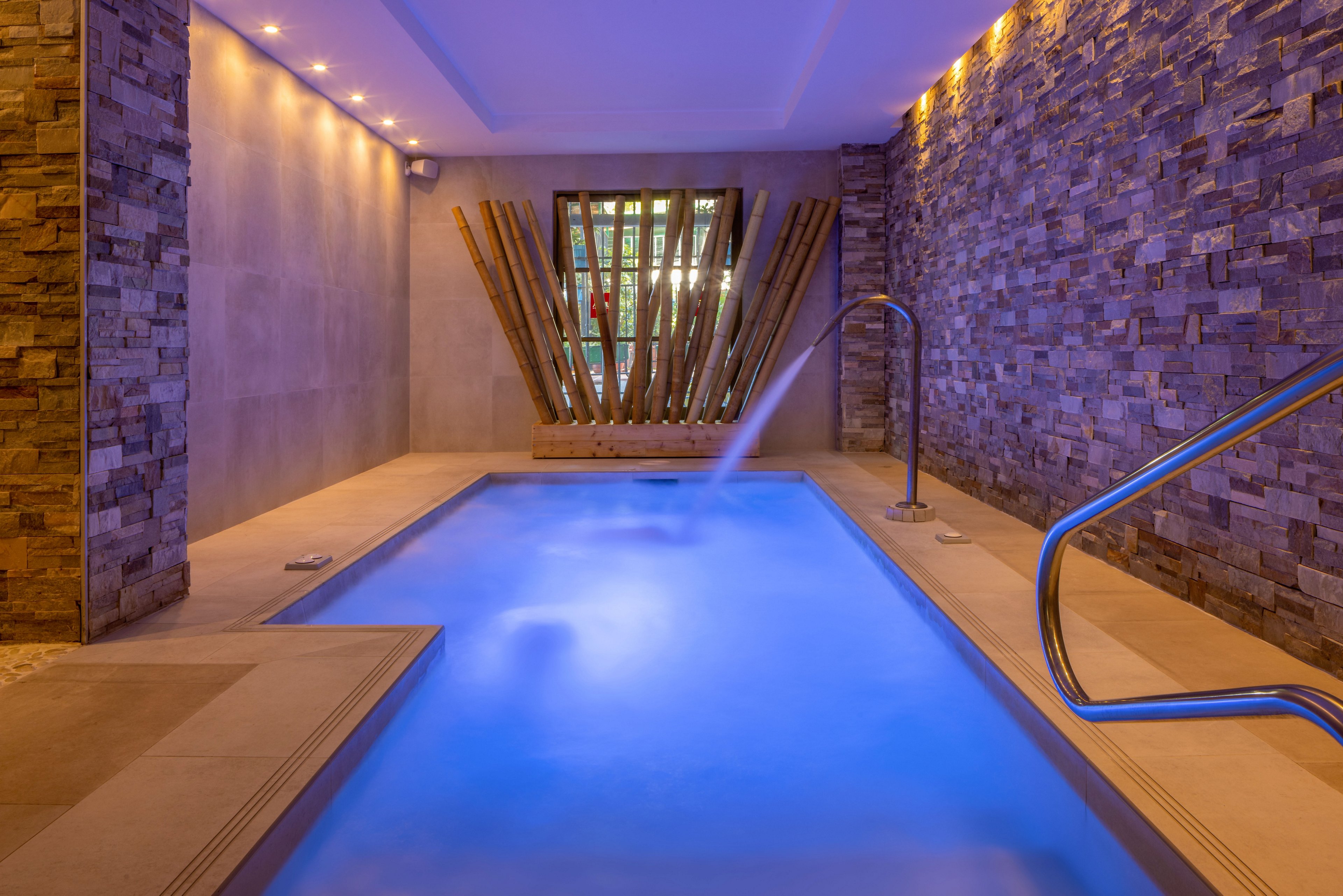 A contemporary indoor spa pool glows with vibrant blue underwater lighting. Stone and concrete walls create a luxurious, minimalist atmosphere. A metal handrail and water jet add functional details, while decorative wooden beams form a striking focal point at the far end. Recessed ceiling lights enhance the warm, relaxing ambiance of the space.