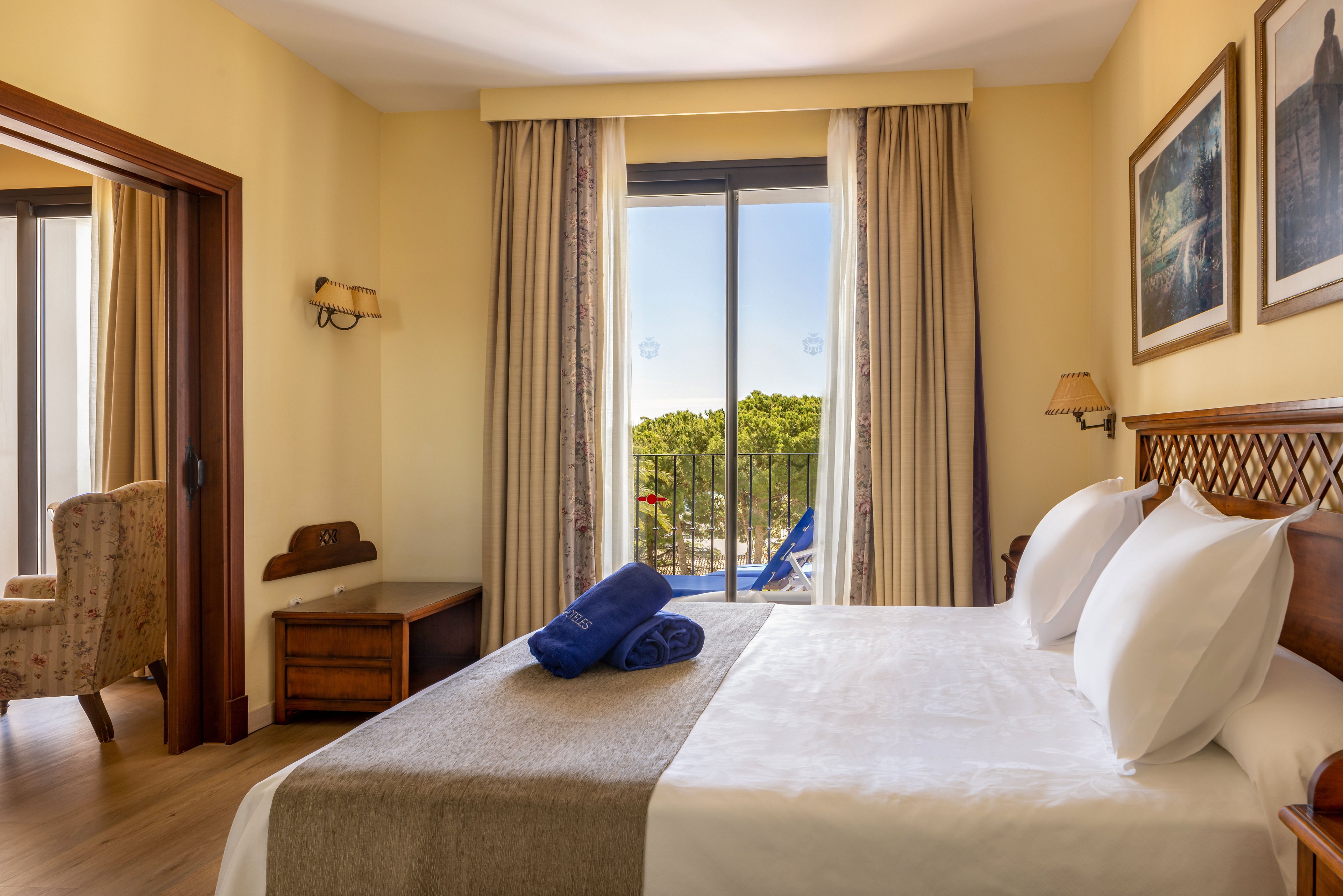 A neatly made double bed with white pillows and a beige throw sits in a bright hotel room. Rolled blue towels rest on the bed, facing a large glass door that opens to a balcony with outdoor chairs and greenery beyond. Warm yellow walls, wooden furniture, and framed artwork create a cozy, inviting atmosphere. An adjacent sitting area with an armchair is visible through an open doorway, enhancing the sense of space and comfort.