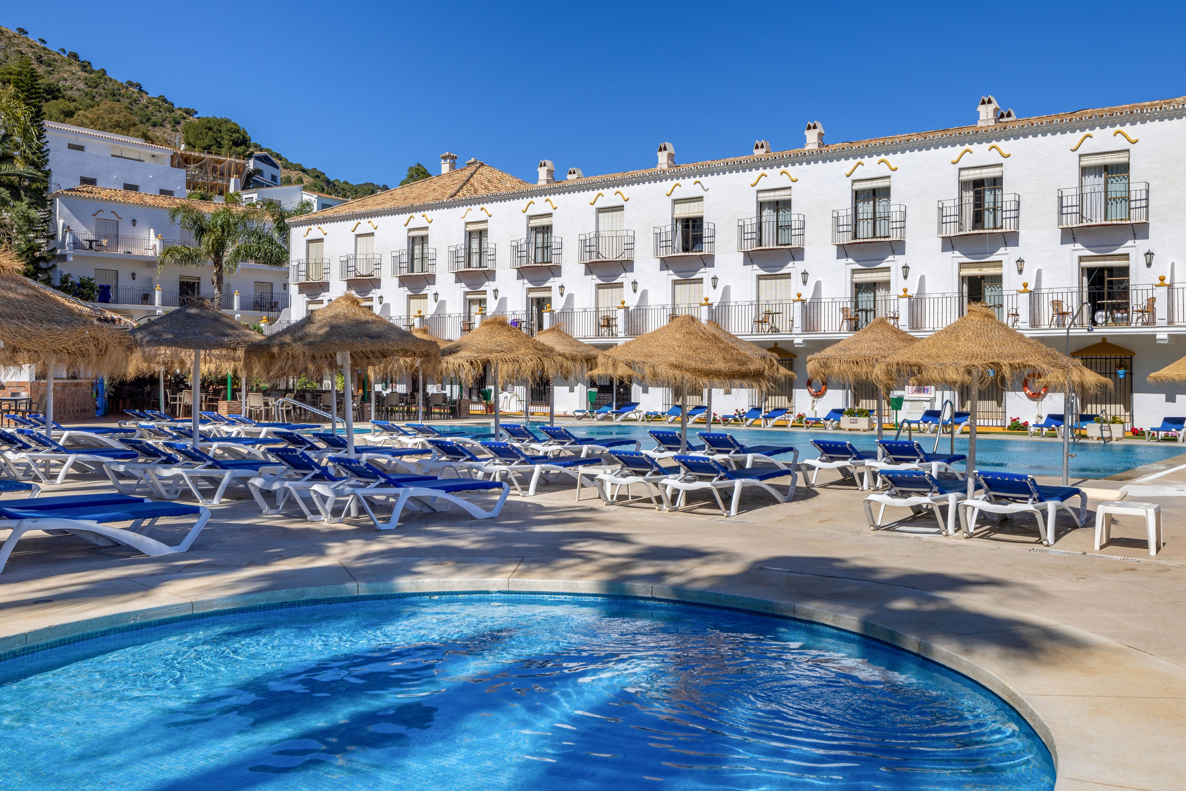 A bright outdoor hotel pool area features a circular shallow pool in the foreground and a larger rectangular swimming pool behind it. Numerous blue sun loungers and straw parasols are arranged neatly around the pools, suggesting a relaxing vacation setting. A white multi-story building with balconies and dark window frames forms the backdrop, with hills and greenery rising behind it under a clear blue sky. The scene conveys a warm, sunny resort atmosphere ideal for leisure and sunbathing.