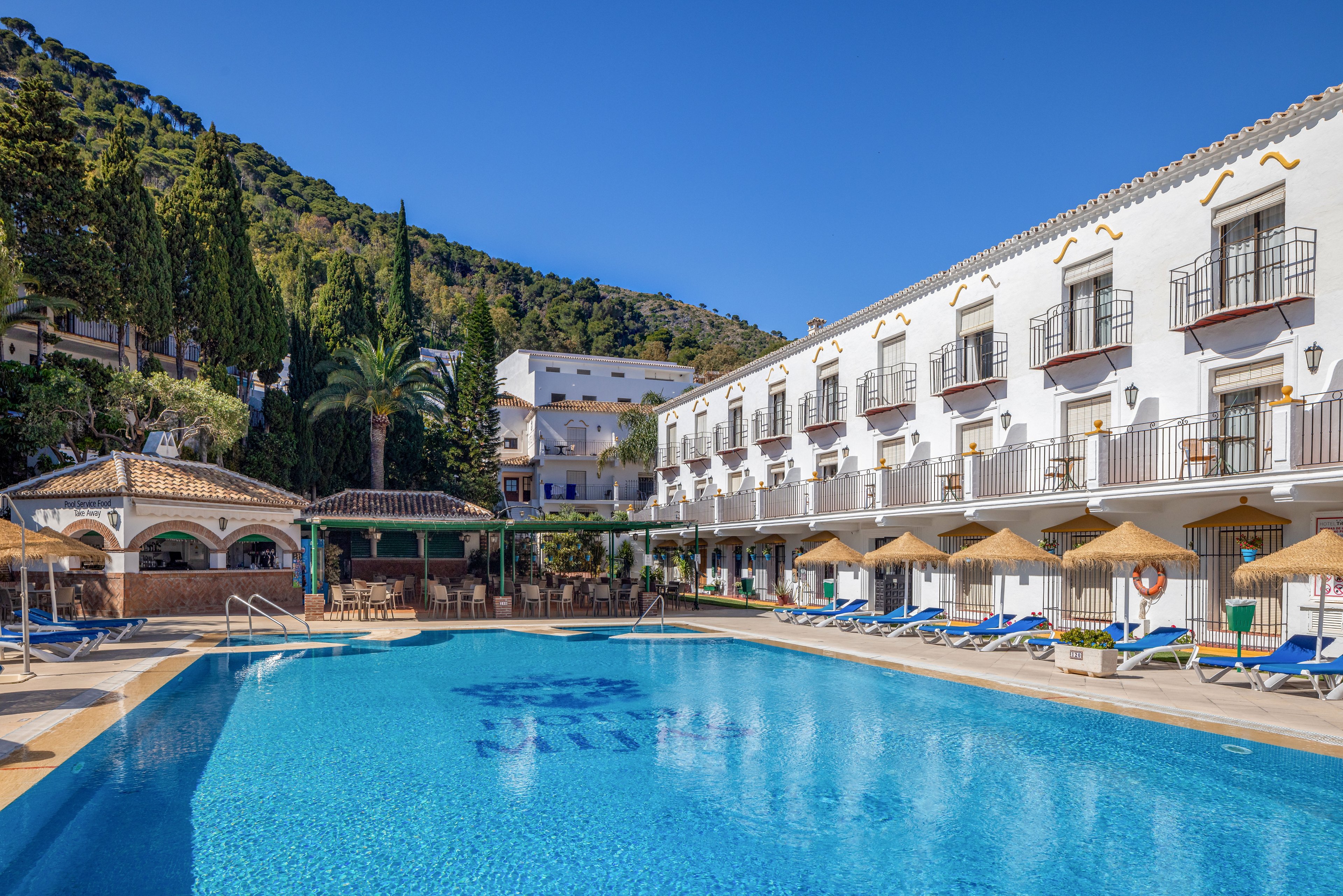 A large outdoor swimming pool sits at the center of a bright hotel courtyard. White multi-story buildings with balconies surround the pool, backed by green hills and trees. Blue sun loungers and straw parasols line the poolside, suggesting a relaxing vacation setting. The clear blue sky and calm water create a warm, inviting atmosphere.