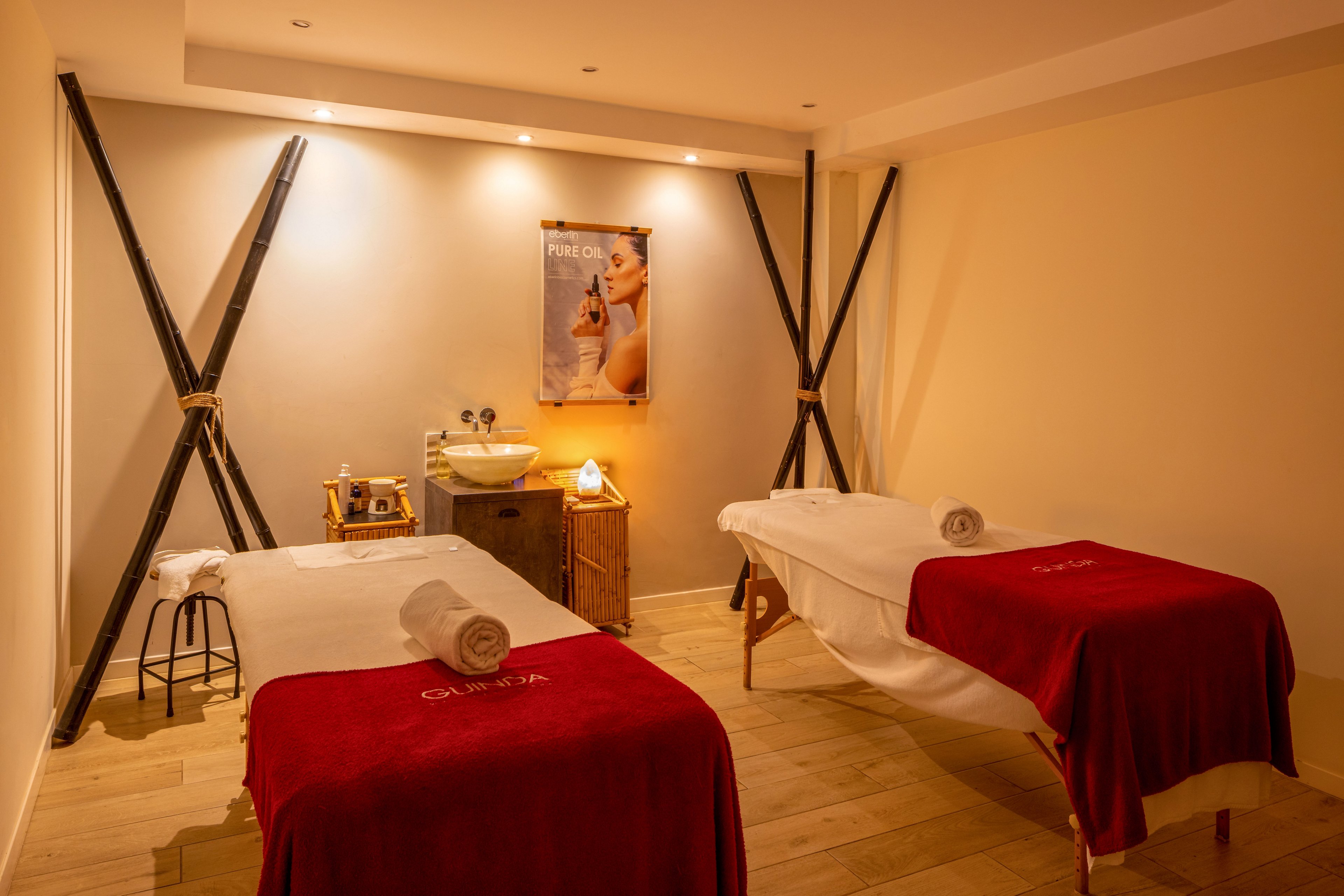 A cozy spa treatment room features two massage tables covered with red blankets and neatly rolled white towels. Soft, warm lighting creates a relaxing atmosphere against light-colored walls and wooden flooring. A small sink area with accessories and a stool sits at the back of the room, framed by decorative dark beams. A wellness poster on the wall adds to the professional spa ambiance.