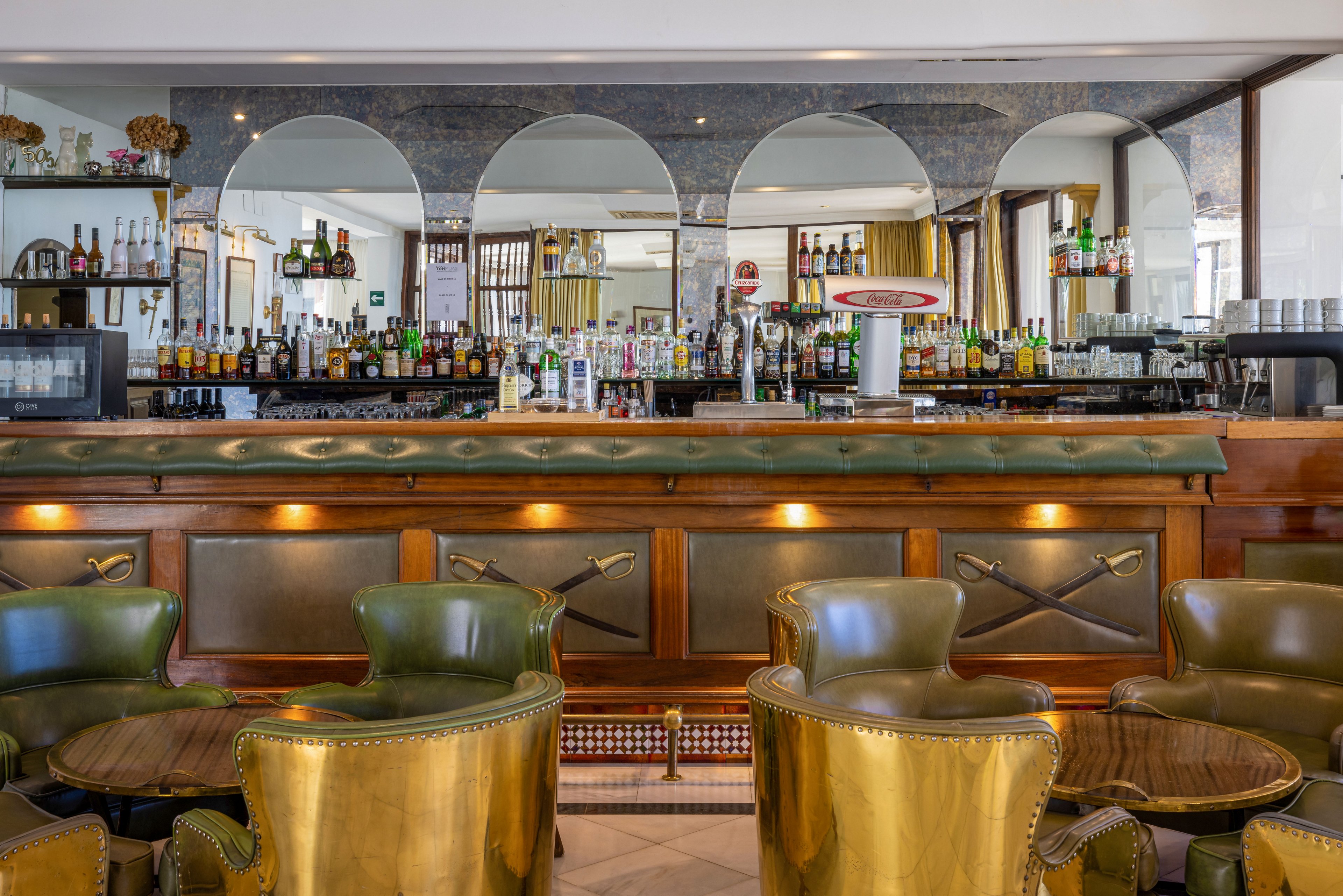 A stylish hotel bar interior features a long wooden counter with green upholstered front panels and brass accents. In front of the bar, several green and gold leather armchairs surround small round tables, creating an intimate seating area. The back bar is lined with assorted bottles, glassware, and bar equipment, reflected in large arched mirrors. Warm lighting under the counter and natural daylight from surrounding windows enhance the sophisticated, vintage atmosphere.