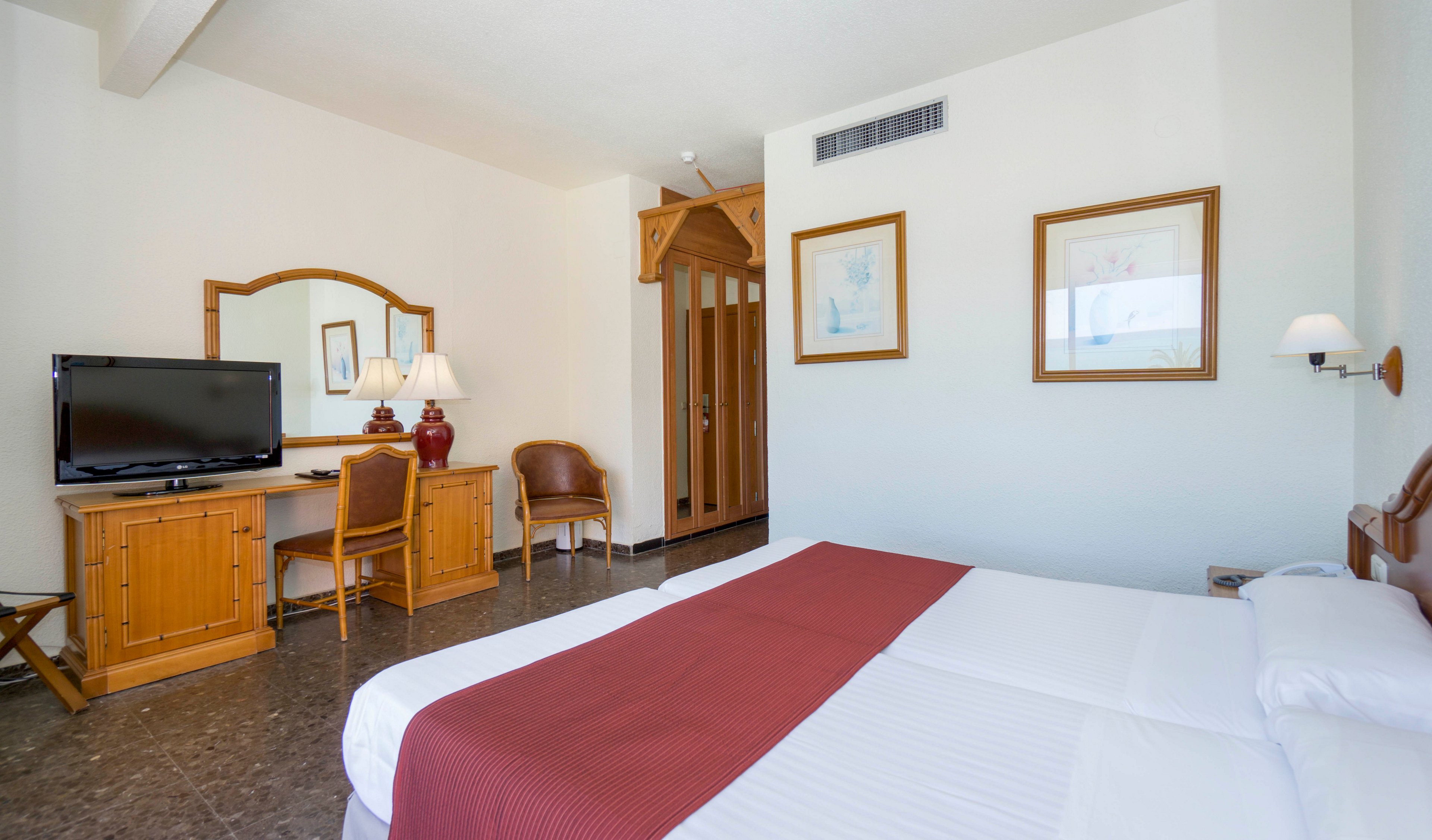 A bright hotel room features a neatly made double bed with white linens and a red runner. Opposite the bed, a flat-screen television sits on a wooden desk with a mirror and table lamp. The room includes a wooden chair, framed wall art, and a small wall-mounted light. Neutral walls and tiled flooring create a clean, simple atmosphere suitable for travelers.
