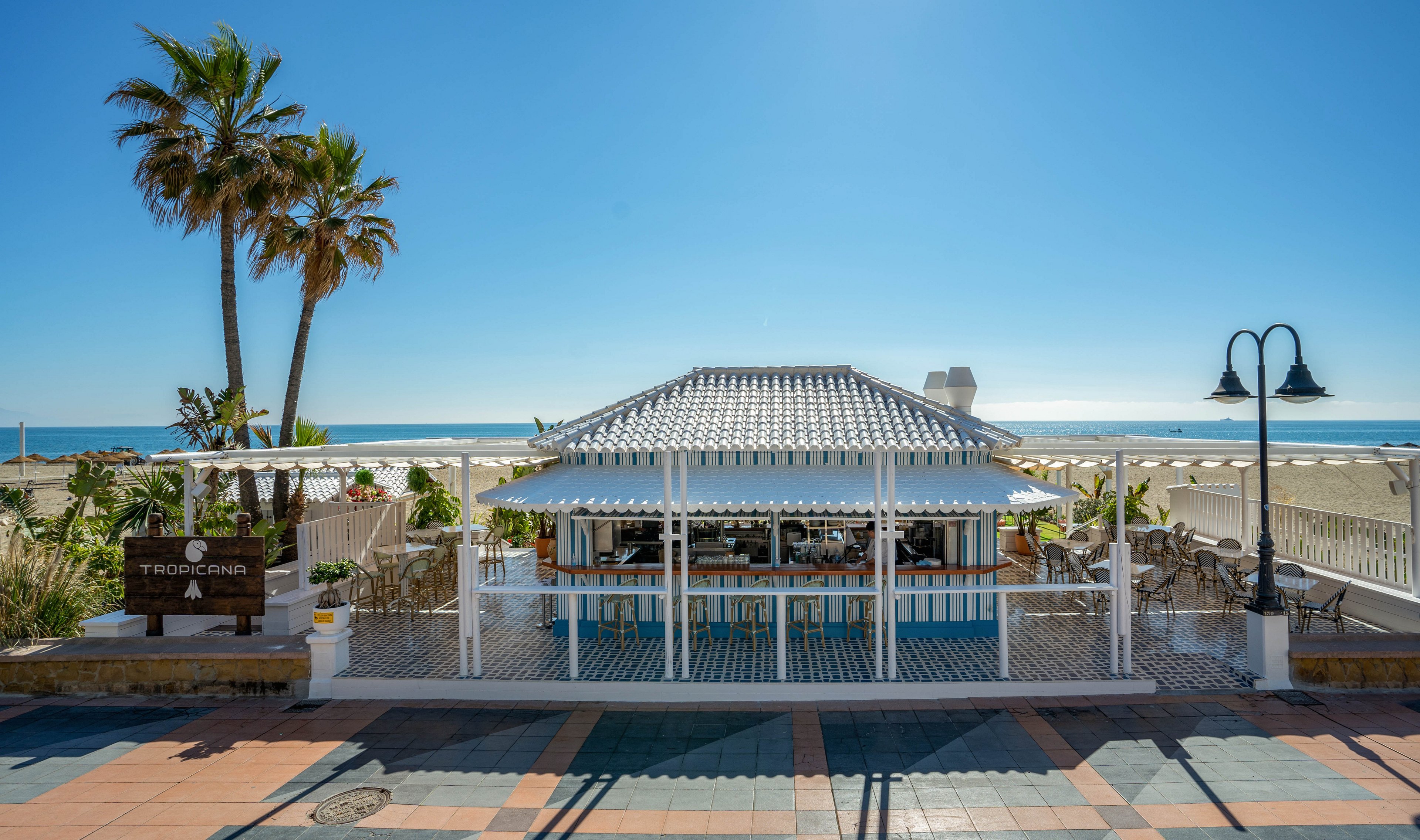 A small open-air bar sits directly on a sandy beachfront under a clear blue sky. White wooden structures and railings frame the bar area, with tables and chairs arranged for outdoor seating. Tall palm trees rise behind the building, and the calm sea stretches out to the horizon. A tiled promenade and street lamps are visible in the foreground, emphasizing the relaxed coastal setting.