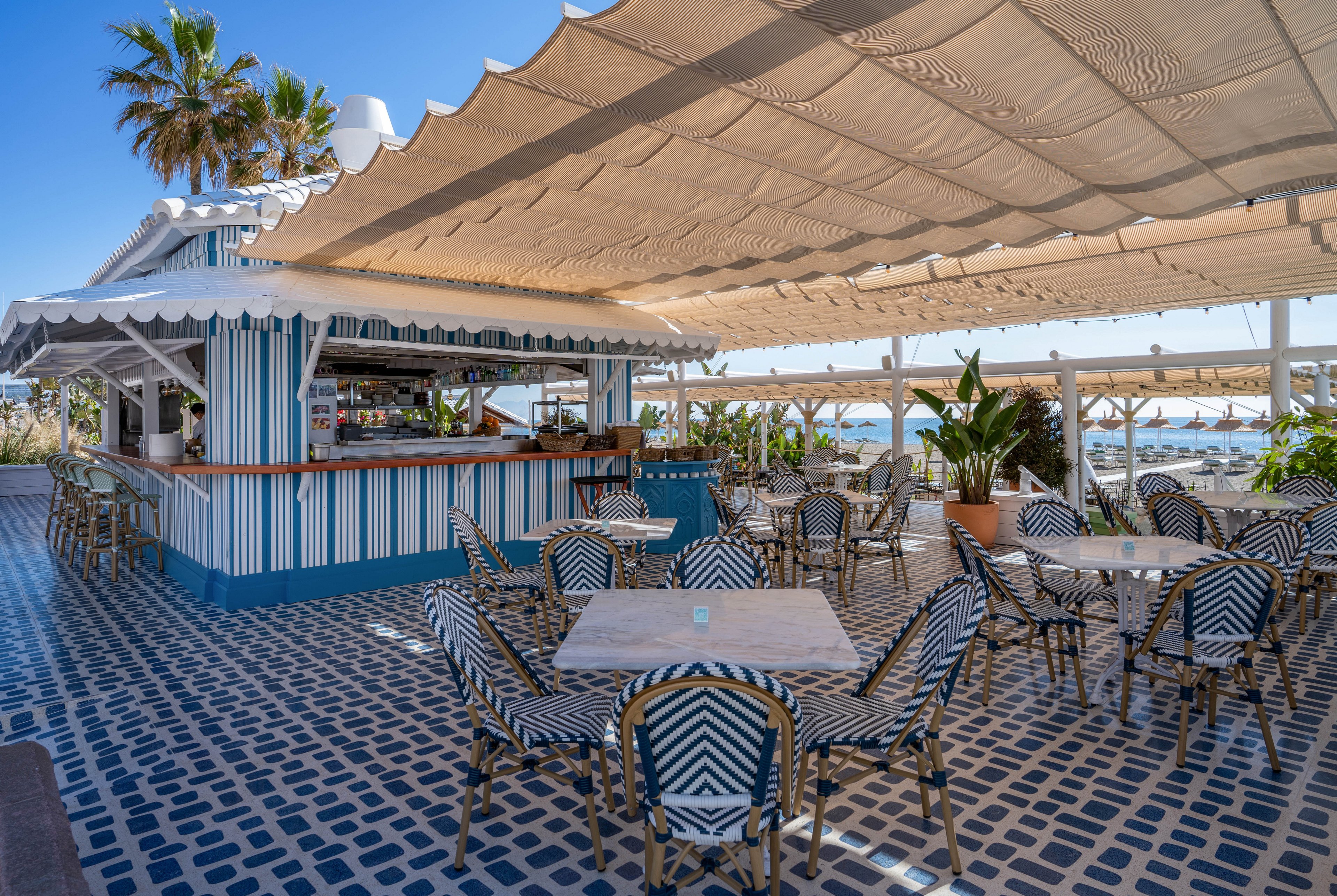 Open-air beach bar terrace featuring striped bar counters and matching chairs under a large canopy. The setting overlooks a sunny seaside with lounge chairs and umbrellas in the background. Patterned tile flooring and potted tropical plants add to the coastal atmosphere. The scene is bright, relaxed, and ready for guests, though no people are currently present.