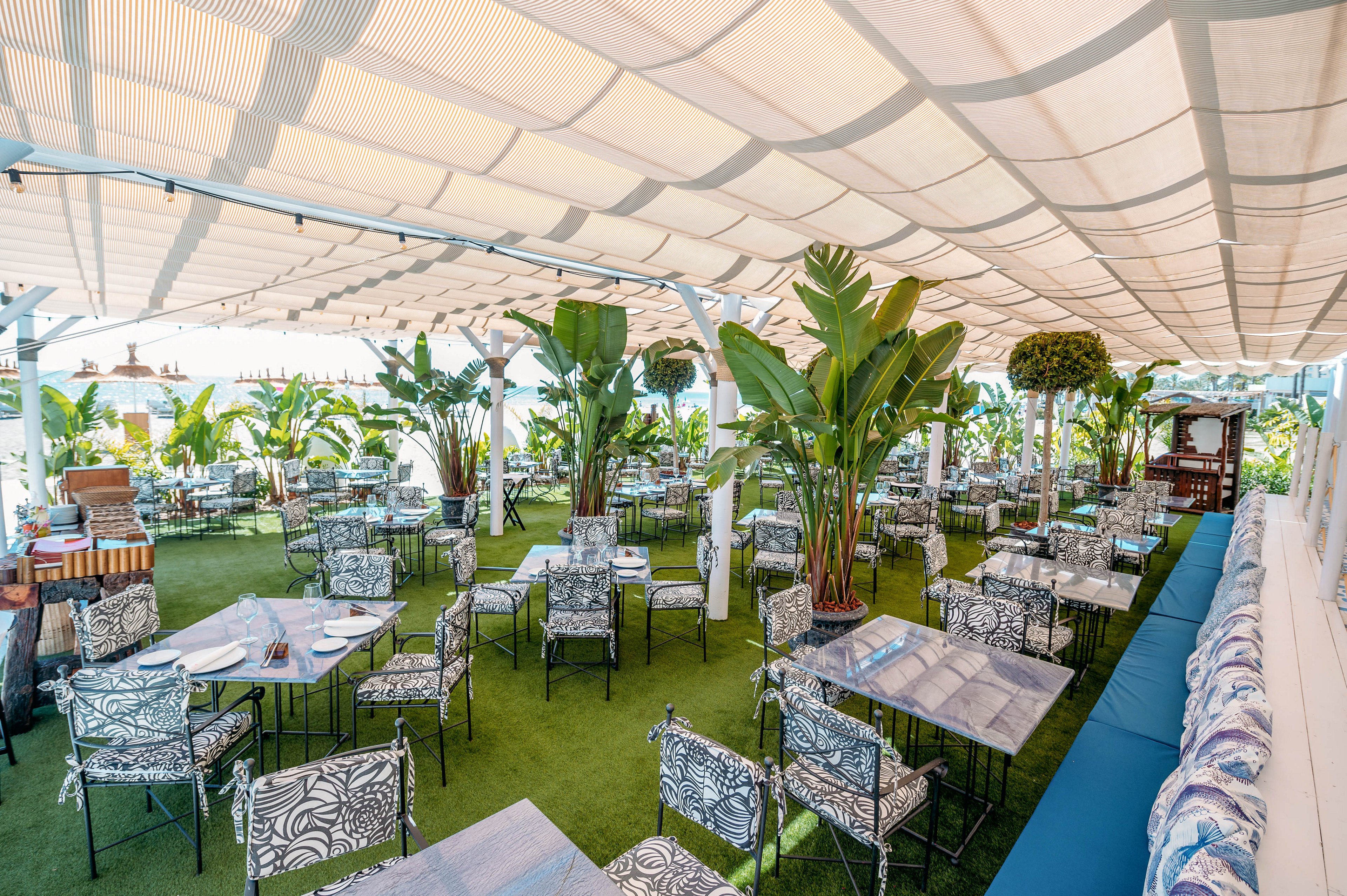 An open-air restaurant features numerous tables and chairs arranged under a large white canopy. The seating area is surrounded by lush tropical plants and greenery, creating a resort-like atmosphere. Patterned cushions and upholstery add decorative detail to the chairs and benches. Natural light filters through the canopy, giving the space a bright, relaxed, and inviting feel.