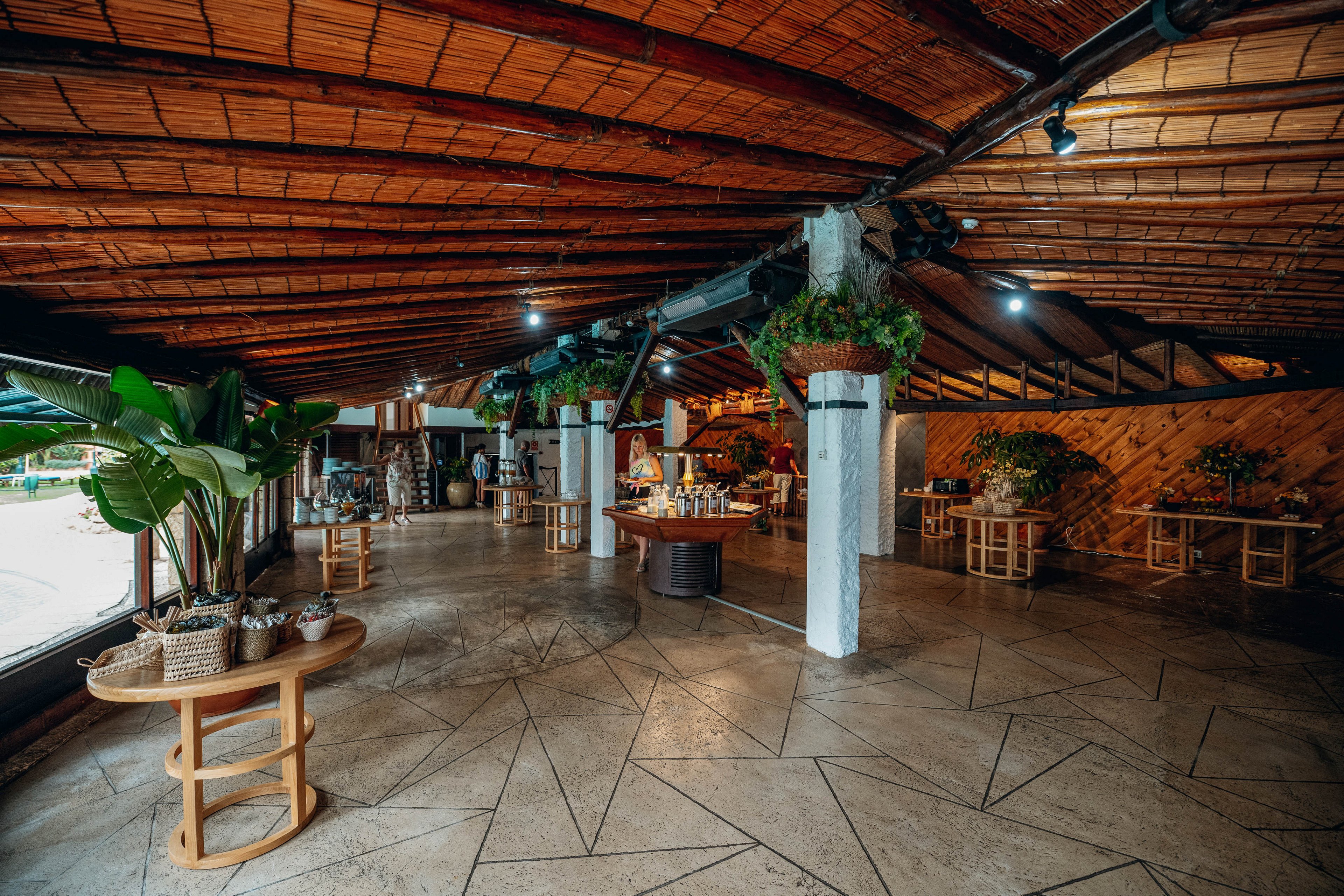 A wide, open interior space features a rustic wooden ceiling and polished stone floor. Several round wooden tables are arranged around the room, some decorated with potted plants and baskets. White columns and hanging greenery add a natural, tropical touch to the setting. The space appears ready for a gathering or event, with warm ambient lighting creating an inviting atmosphere.