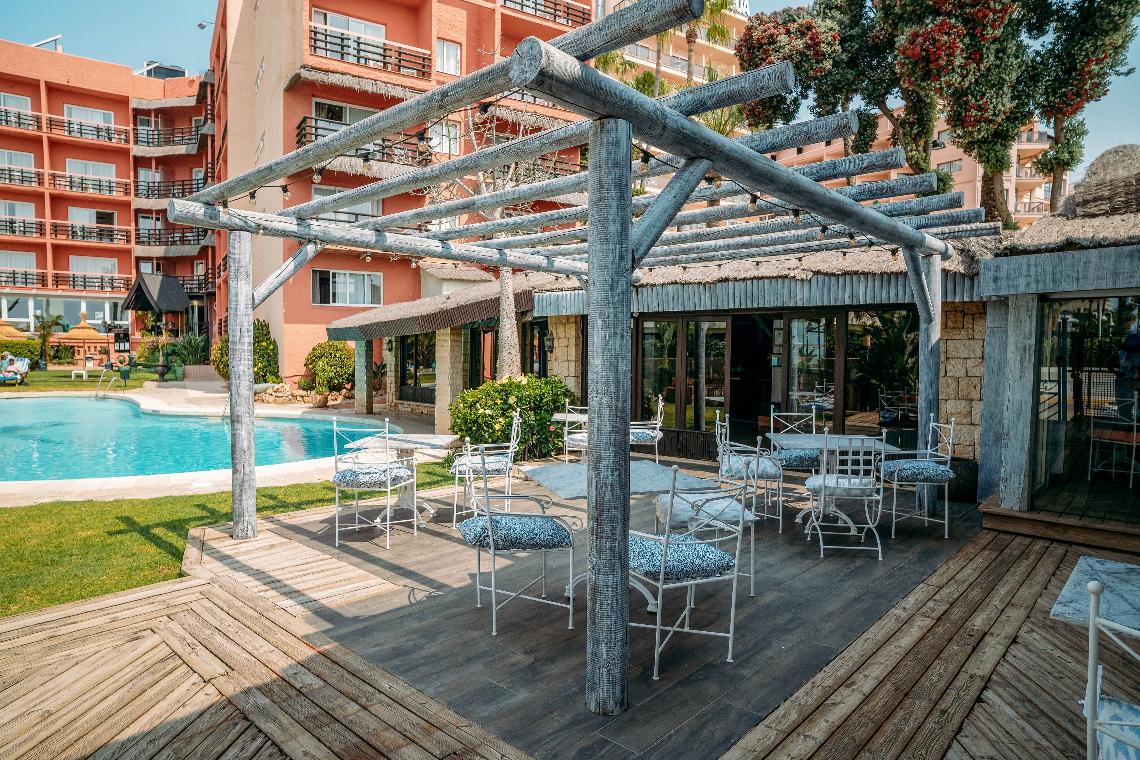 An outdoor hotel terrace features white metal chairs and cushioned seats beneath a wooden pergola. The seating area overlooks a clear blue swimming pool surrounded by grass and wooden decking. A multi-story building with balconies and large windows forms the backdrop. The scene suggests a relaxed resort atmosphere on a sunny day.