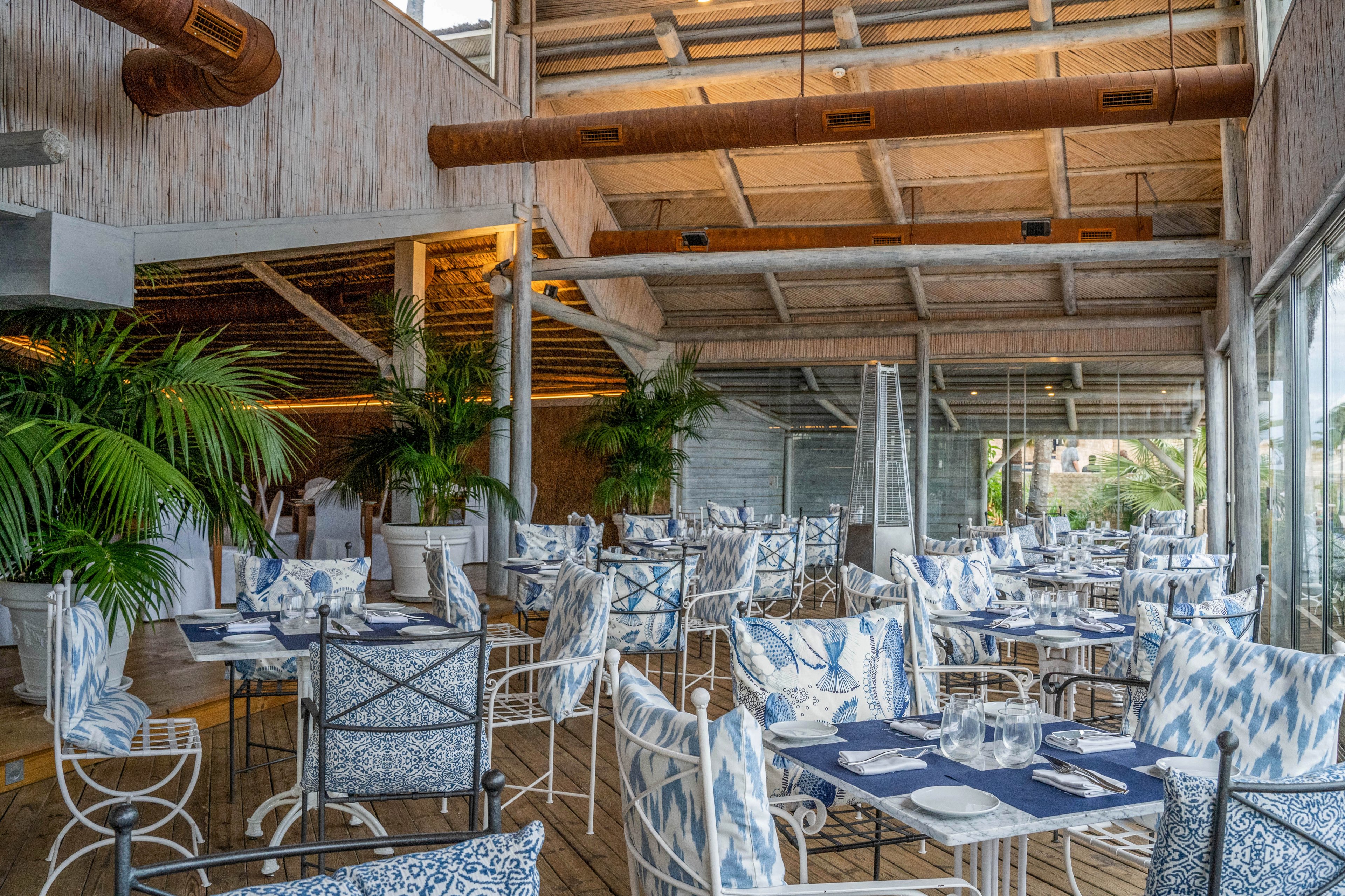 Bright coastal restaurant interior with neatly arranged dining tables and chairs. The space features large windows, wooden ceilings, and exposed beams creating an airy, open atmosphere. Blue and white patterned cushions and table linens add a nautical touch to the decor. Potted palm plants and natural light enhance the relaxed, beachside ambiance.