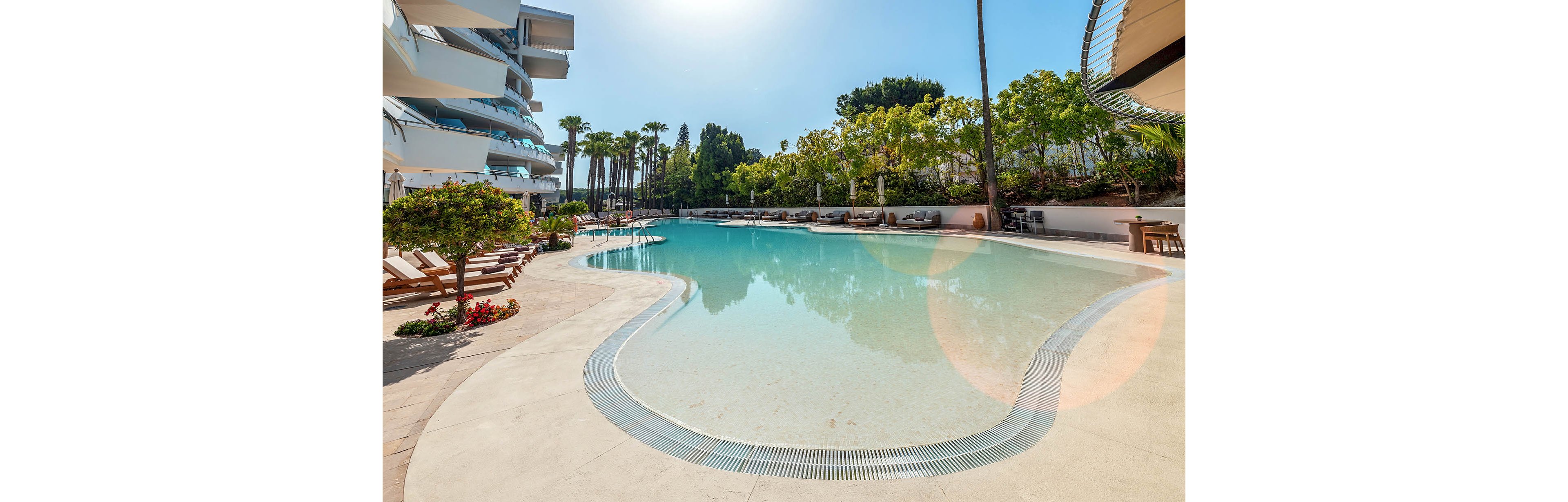 A large, curved swimming pool is shown in a modern resort setting under bright sunlight. Sun loungers line the poolside terrace, some shaded by trees and umbrellas, creating a relaxing vacation atmosphere. Contemporary white buildings and tall palm trees frame the scene, reflecting in the calm turquoise water. The overall mood is tranquil and luxurious, ideal for leisure and summer holidays.