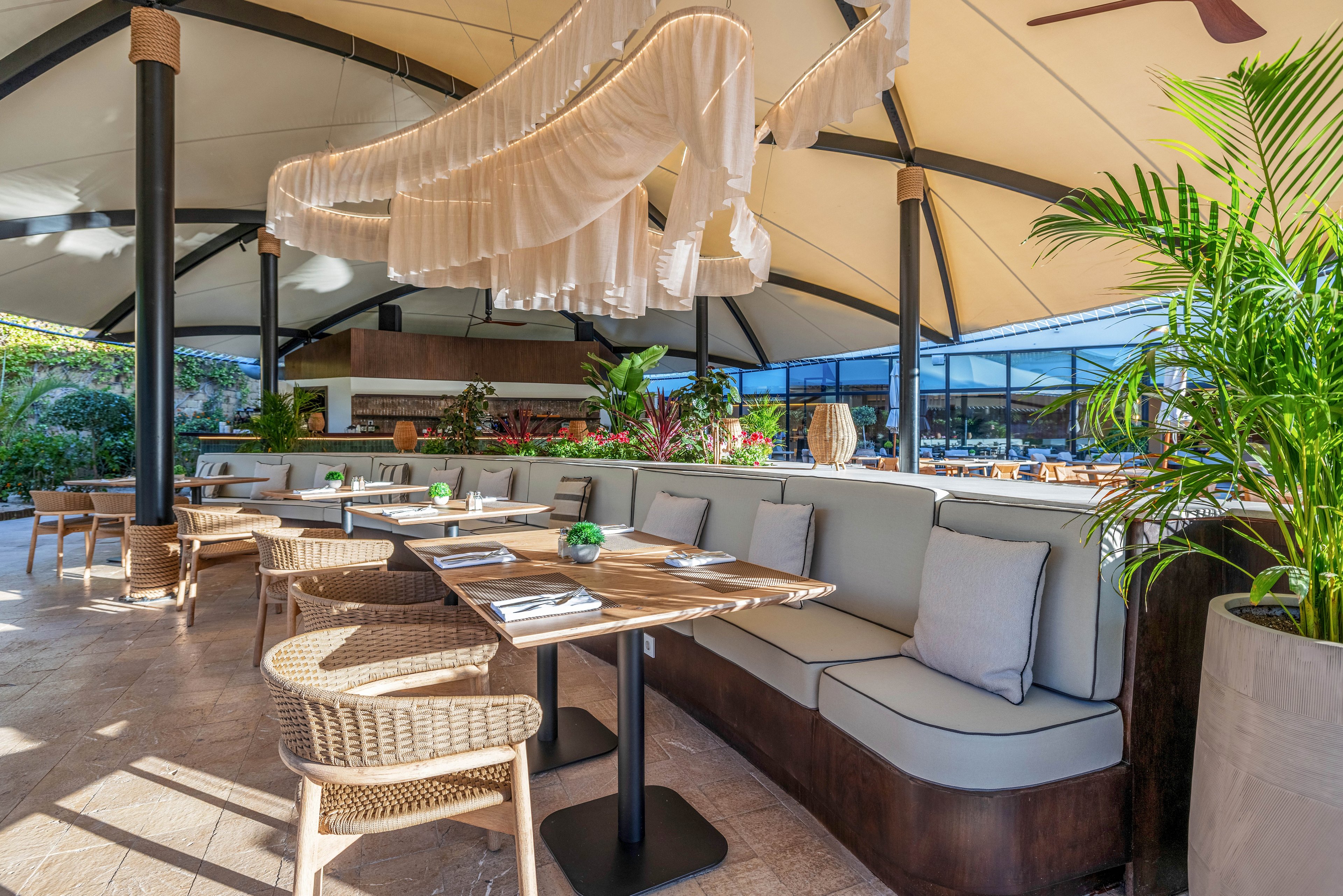 Bright outdoor restaurant terrace featuring a long cushioned banquette and wooden tables set for dining. Wicker chairs and neutral upholstery create a relaxed, contemporary atmosphere under a large canopy roof. Decorative hanging fabric elements and lush green plants add visual interest and a resort-like feel. Sunlight floods the open space, highlighting the clean, inviting setup ready for guests.