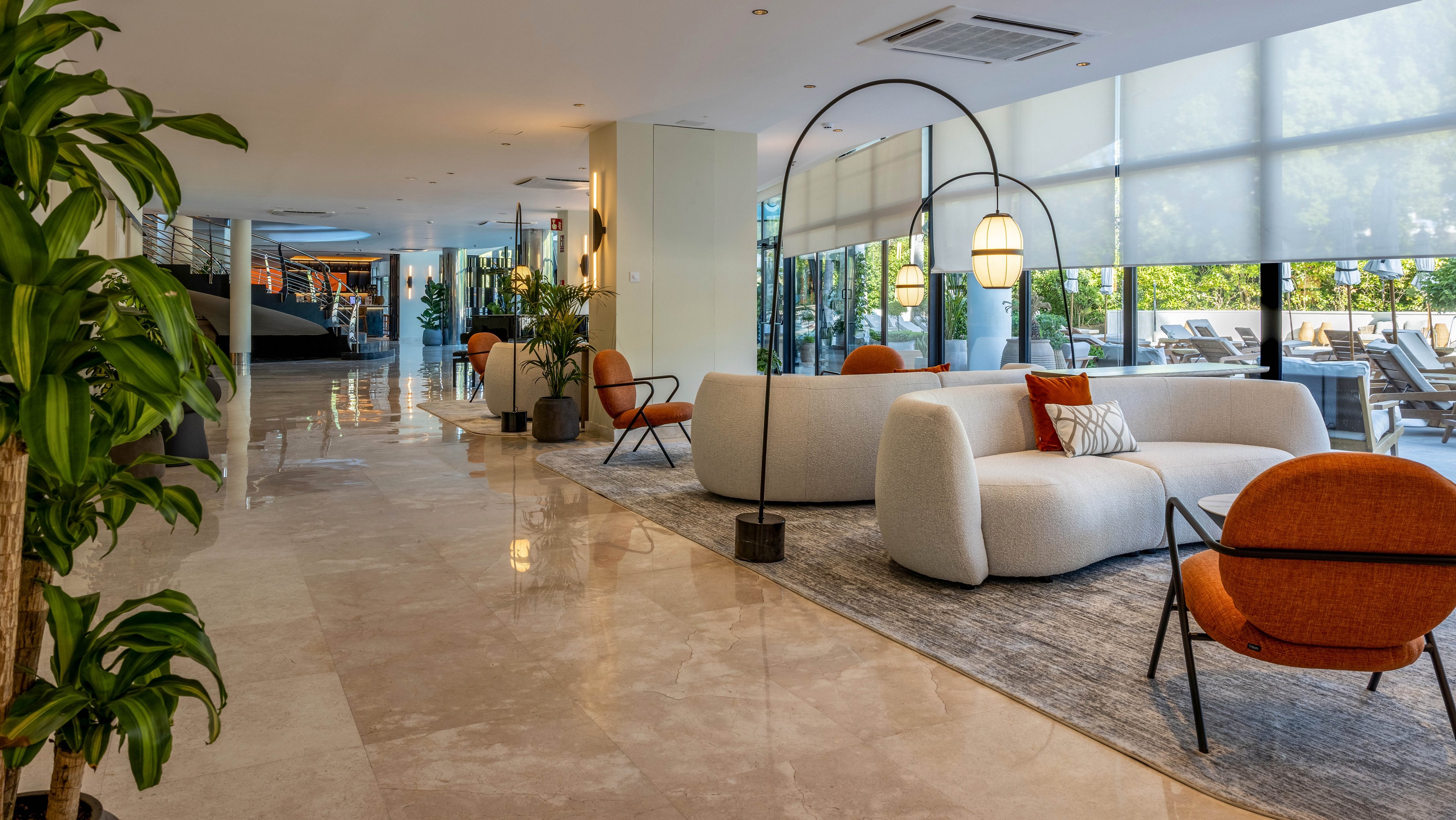 A spacious, modern lobby features curved white sofas and orange accent chairs arranged on a large area rug. Tall arched floor lamps with hanging lights create a cozy seating zone along the polished stone floor. Large windows with sheer blinds let in natural daylight and reveal an outdoor terrace with loungers. Green potted plants add a touch of nature to the clean, minimalist interior design.