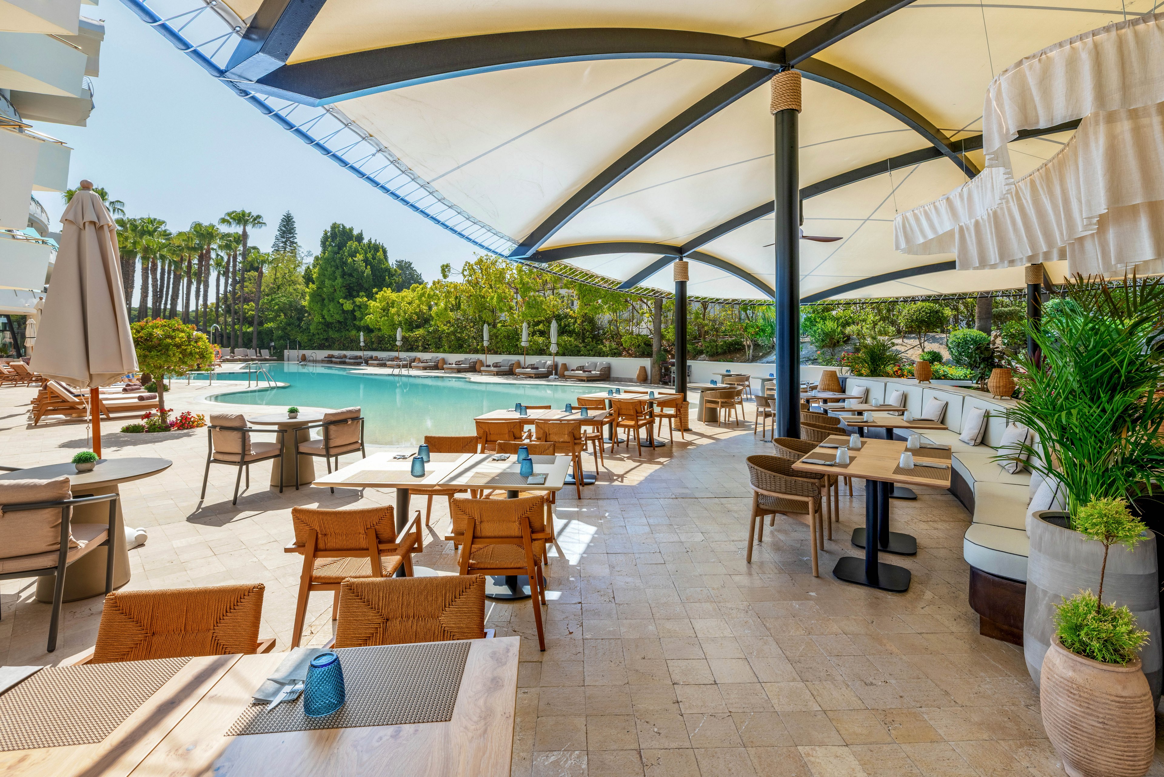 An open-air restaurant terrace overlooks a bright turquoise swimming pool at a modern resort. Wooden tables and chairs are neatly arranged under large white canopy structures providing shade. Cushioned bench seating lines one side, complemented by potted green plants and neutral stone flooring. The scene is sunlit and inviting, suggesting a relaxed vacation atmosphere in a warm climate.