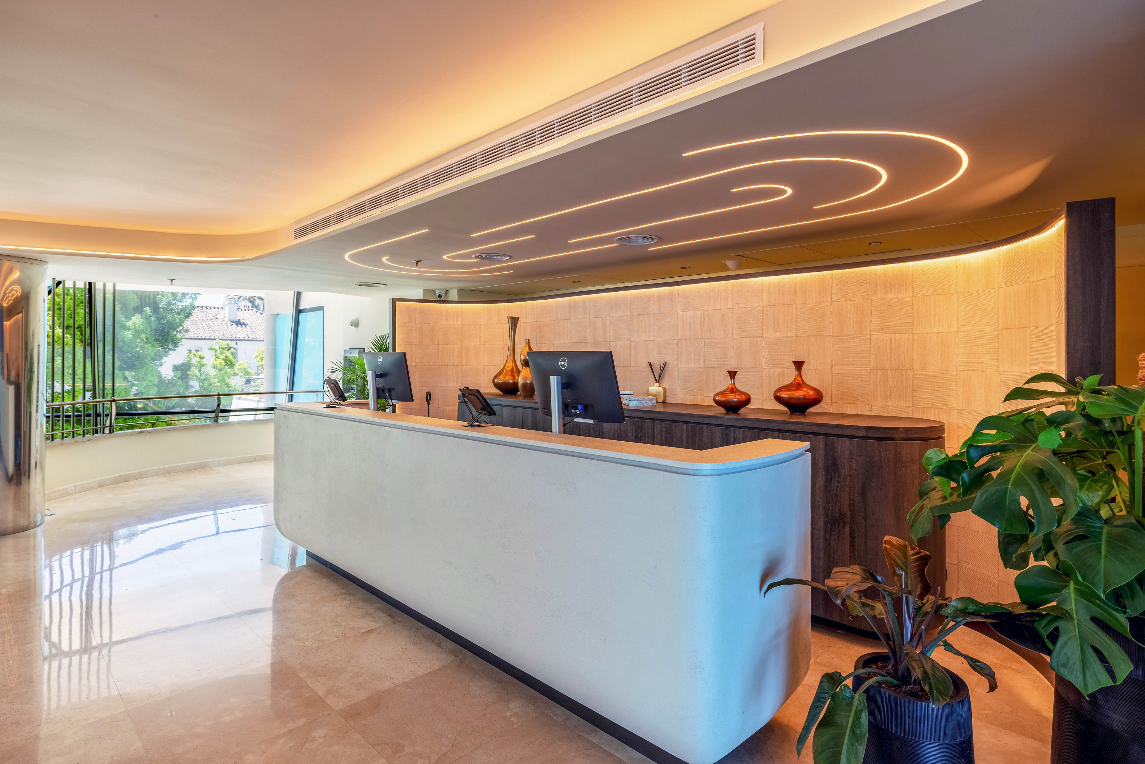 A contemporary reception desk stands in a bright hotel lobby with smooth, curved surfaces. Warm ceiling lights create an elegant pattern above the counter, complementing the neutral stone floor. Computer monitors and decorative vases sit on the dark wood counter, adding functional and stylish elements. Large windows and indoor plants bring natural light and greenery into the space, enhancing the welcoming atmosphere.