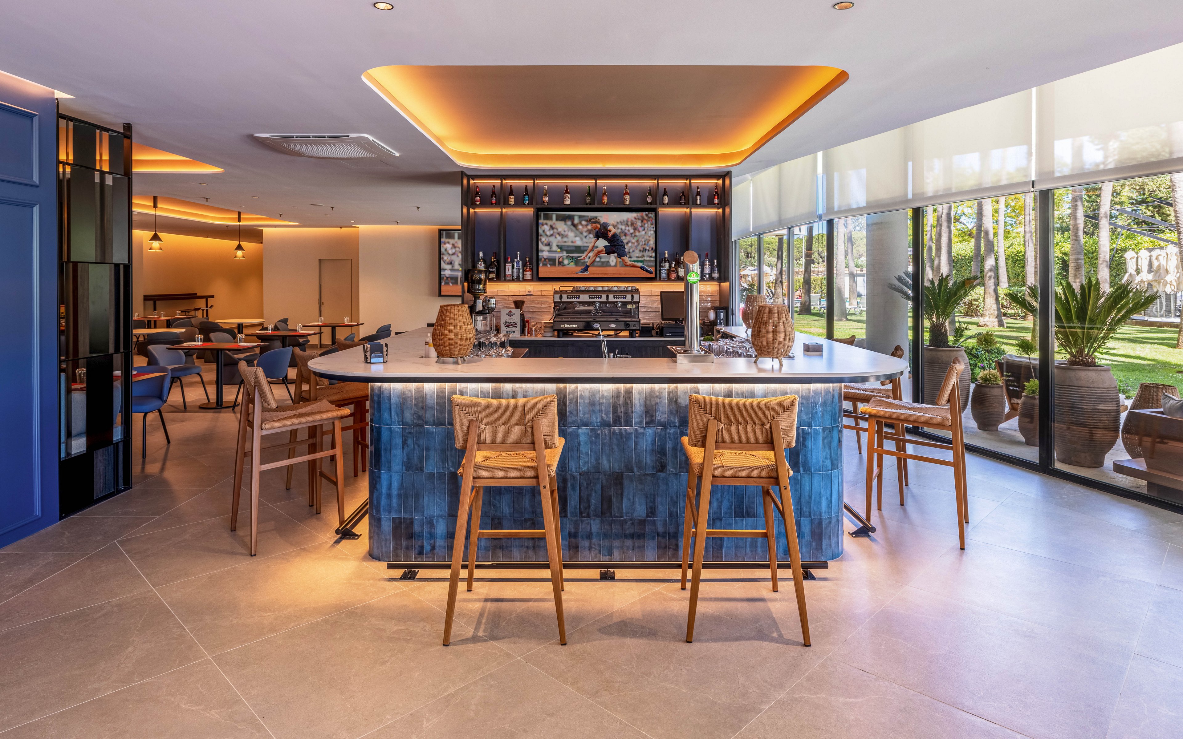 A contemporary bar with a central counter and wooden stools sits in a bright lounge area. Warm ceiling lighting and pendant lamps create an inviting atmosphere around the bar. Large windows reveal a landscaped garden with palm trees and decorative barrels outside. The space features clean lines, neutral flooring, and blue accents for a stylish, modern look.