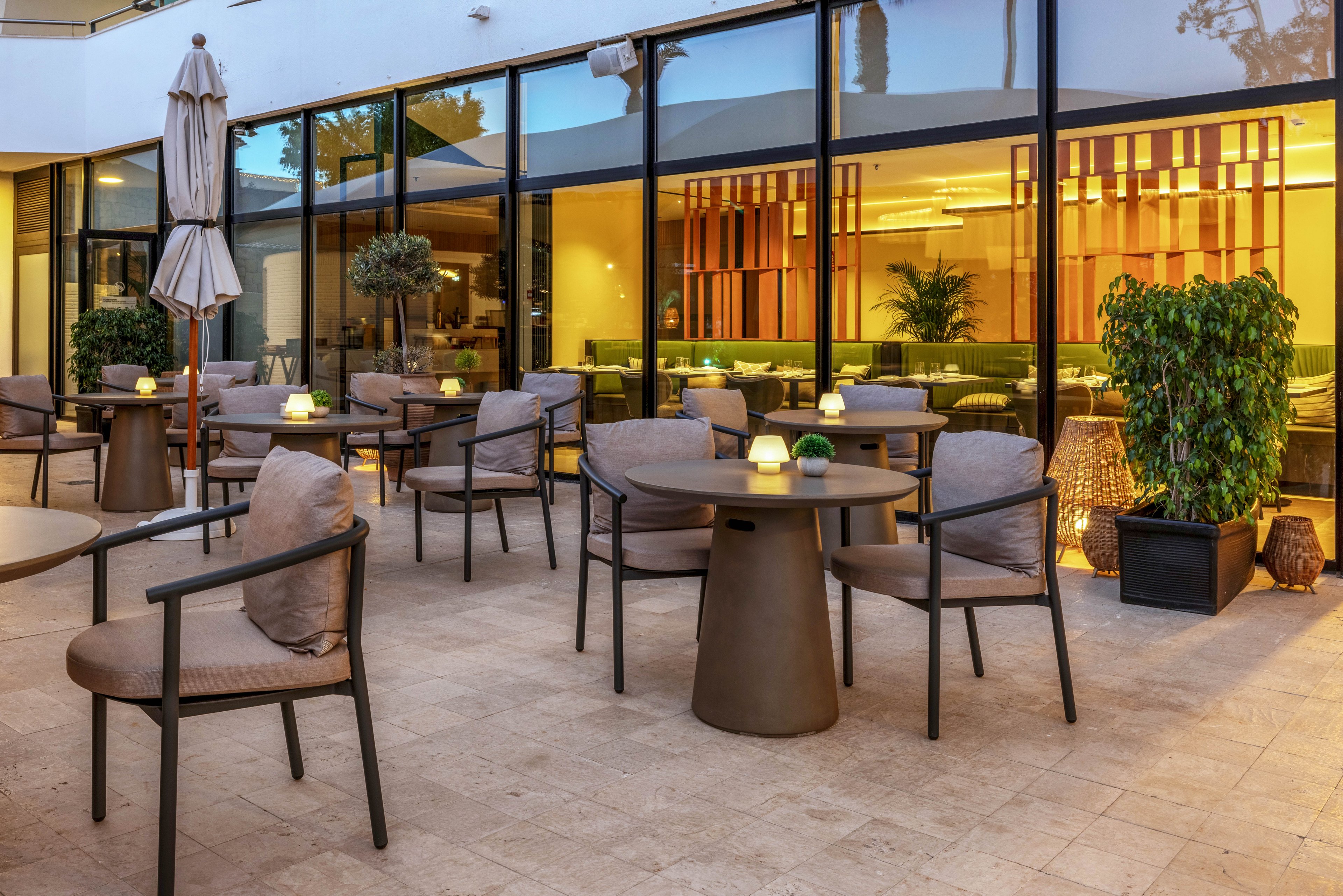 Contemporary outdoor patio area with multiple round tables and cushioned chairs is shown at dusk. Small candle-like lights sit on the tables, creating a warm and inviting atmosphere. Large glass windows reveal an indoor lounge with soft yellow lighting and modern decor. Potted plants and closed umbrellas add to the relaxed, upscale setting.