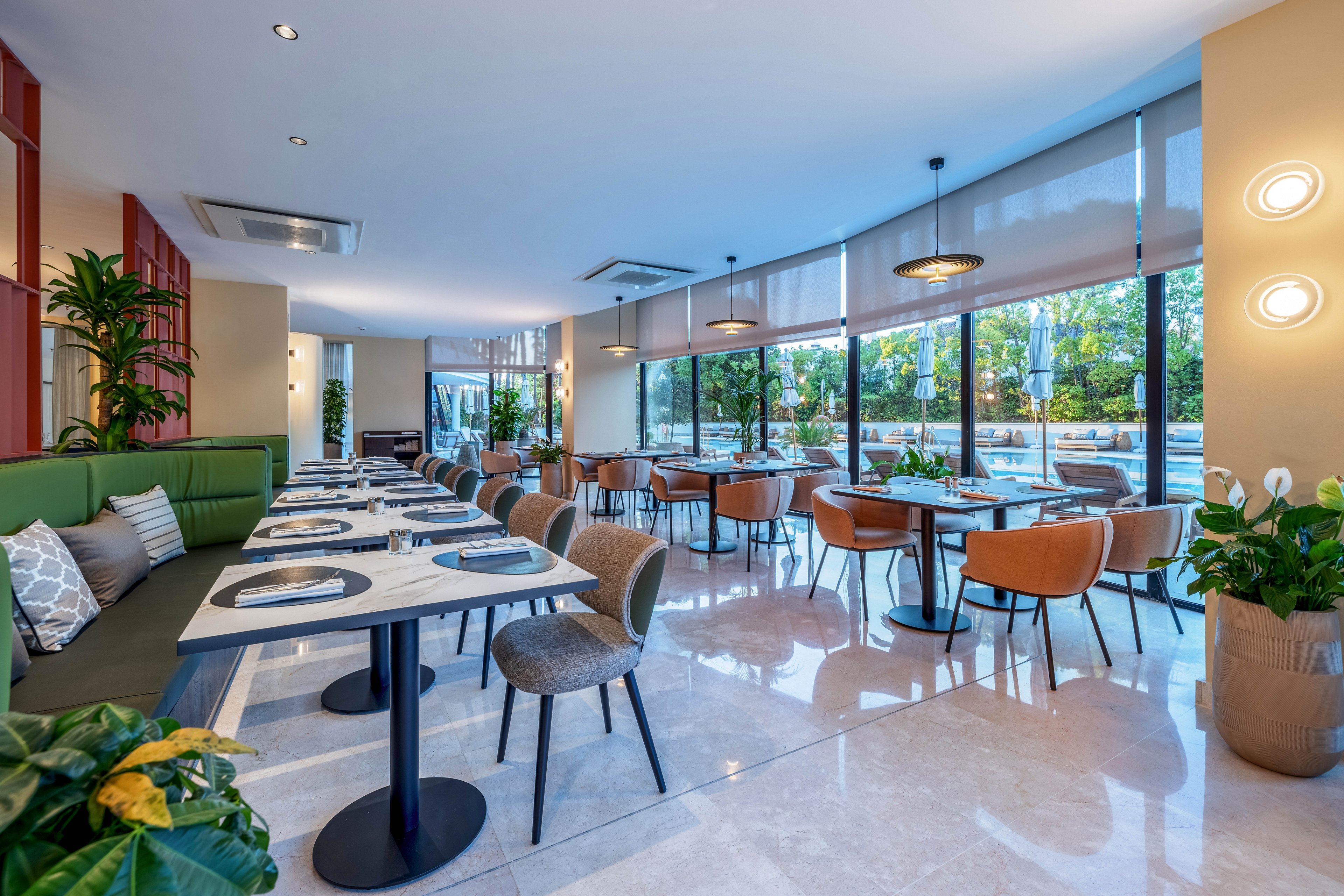 Contemporary restaurant dining area featuring neatly arranged tables and chairs. Large floor-to-ceiling windows let in abundant natural light and offer views of greenery outside. A long upholstered bench with cushions lines one side, while pendant lights and wall sconces add warm illumination. Polished flooring and indoor plants contribute to the clean, inviting atmosphere.
