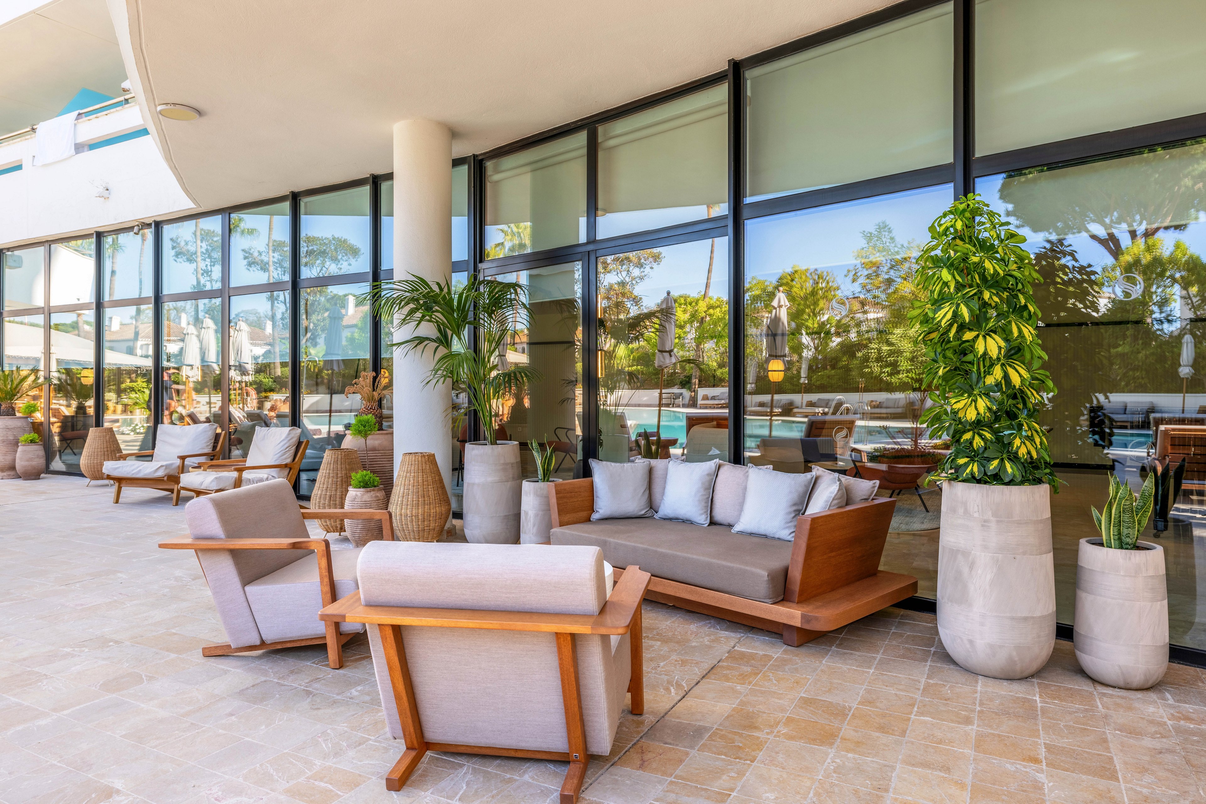 Contemporary wooden lounge seating is arranged on a spacious outdoor terrace beside large glass windows. Cushioned sofas and armchairs create a comfortable relaxation area overlooking a pool and trees. Tall potted plants and wicker accents add natural texture and greenery to the bright, sunlit space. The clean architectural lines and neutral tones convey a modern, resort-like atmosphere.